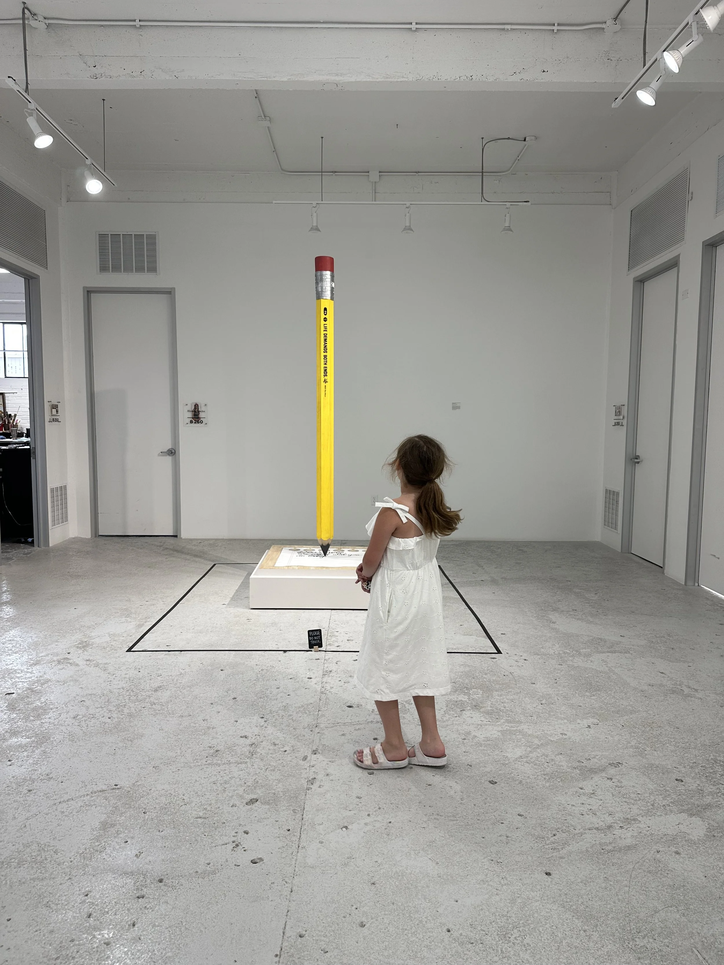 A young girl in a white dress and sandals stands in front of an art exhibit featuring a large yellow pencil sculpture pointing downward, with the girl observing it.