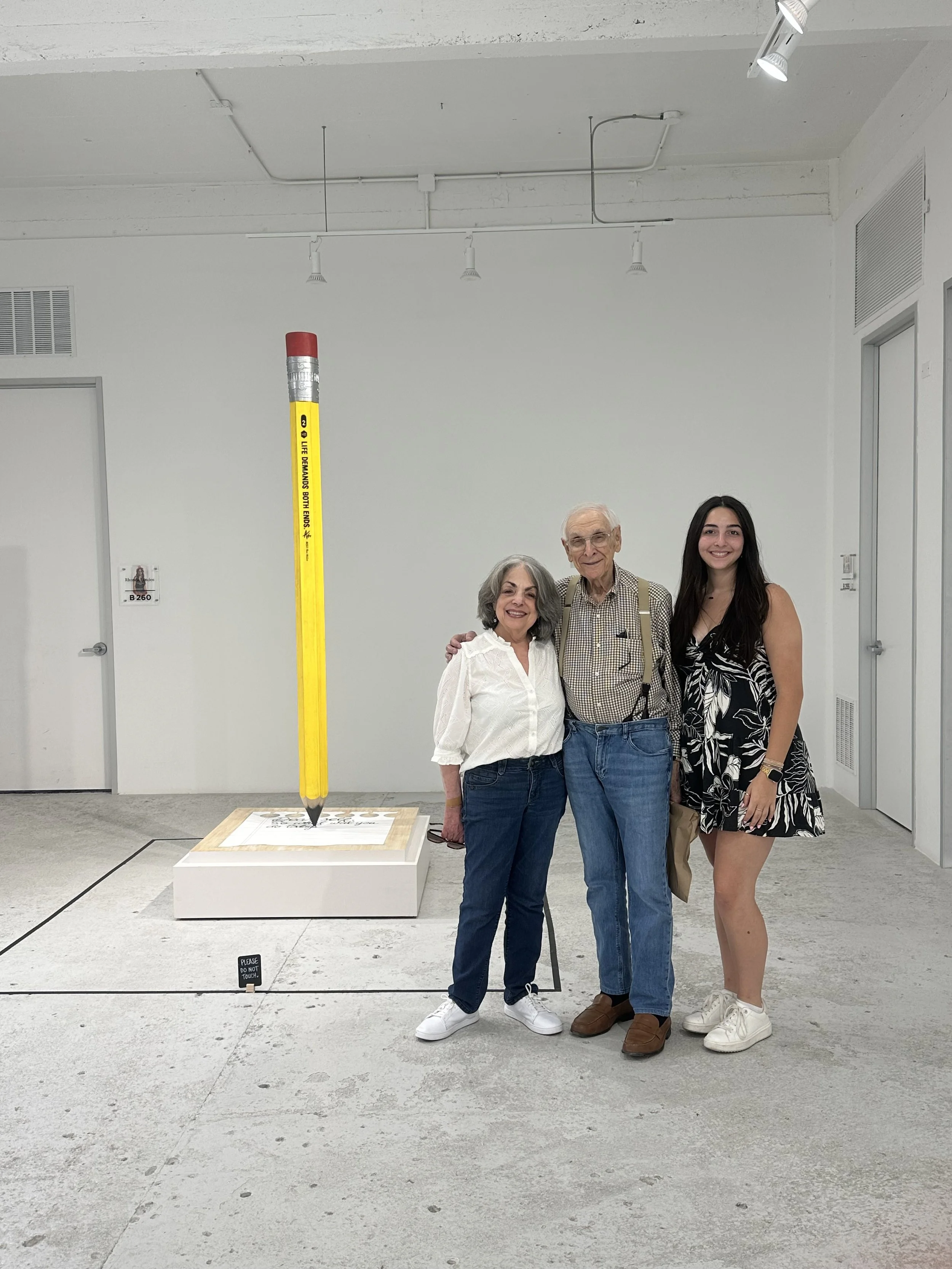 Three people standing together in an art gallery with a large pencil sculpture on display behind them. The group consists of an elderly woman, an elderly man with glasses, and a young woman. They are smiling and posing for the photo.