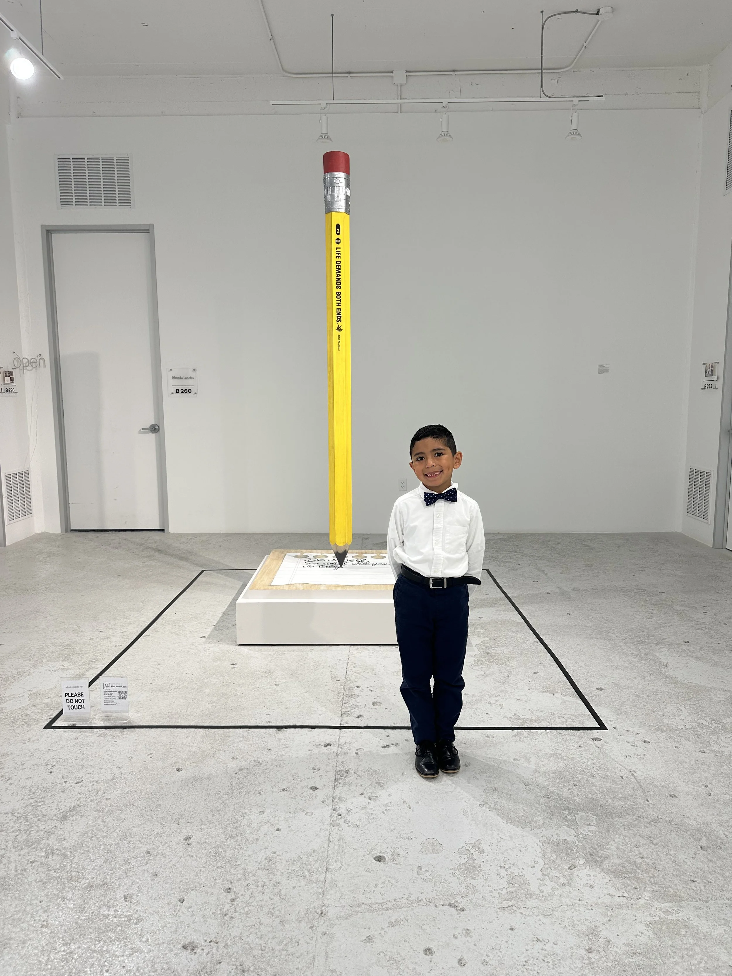 Young boy in white shirt, navy pants, and bow tie standing in front of an art installation of a giant yellow pencil with a red top, with a sign requesting not to touch.