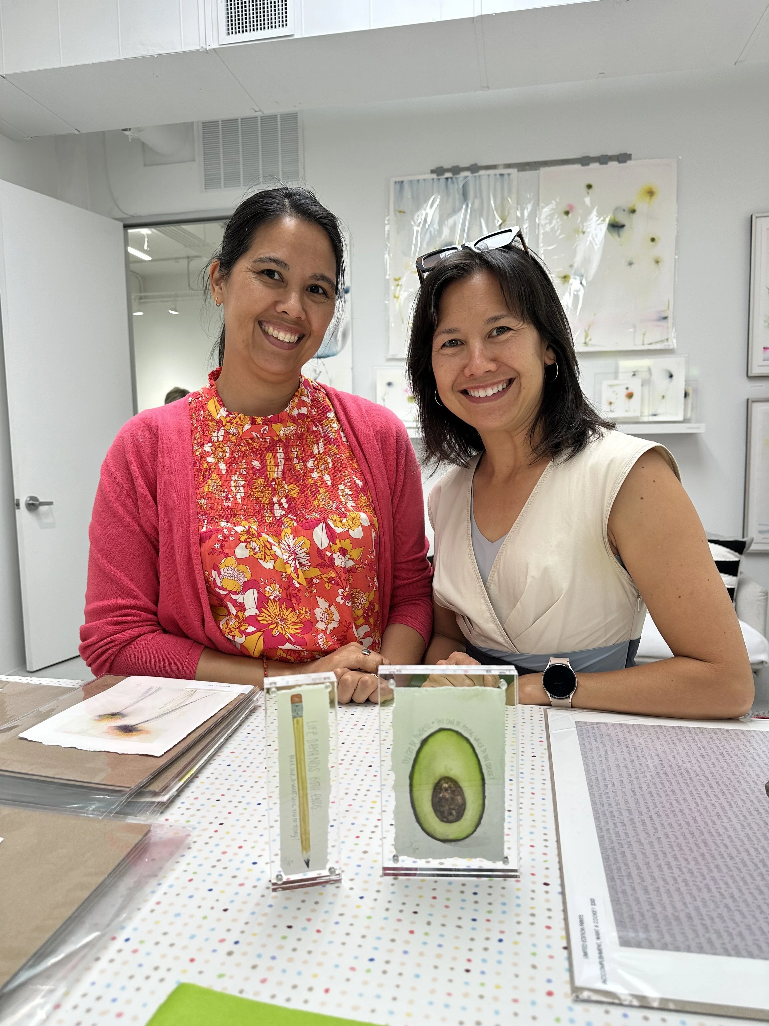 Two women smiling and standing behind a table with art displays, in an art gallery or studio.
