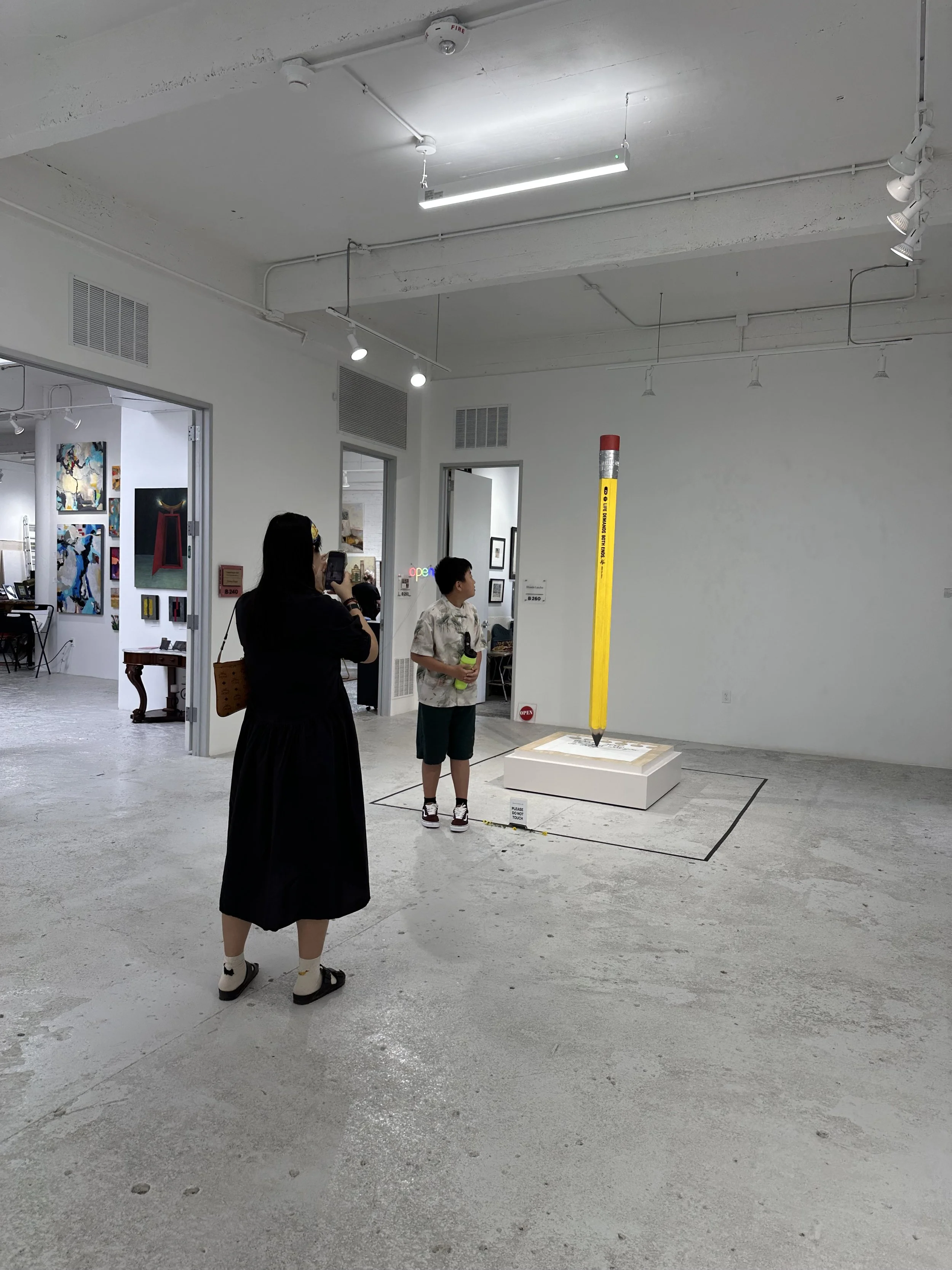 Art gallery with people viewing a sculpture of a giant yellow pencil with a red eraser on top, displayed on a white platform in a white-walled room.