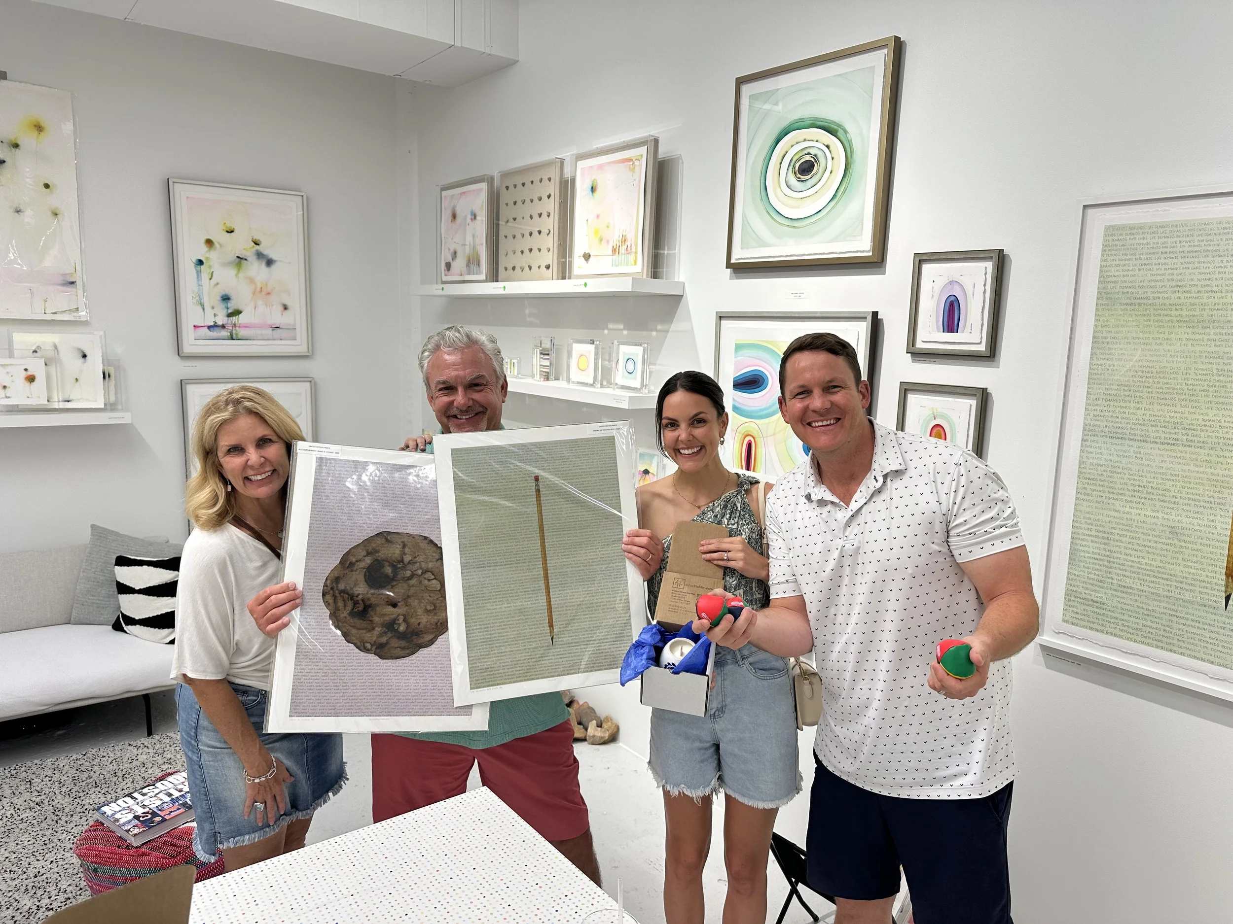 Four smiling people in an art gallery holding up artwork and small balls, standing in front of abstract colorful paintings on the wall.