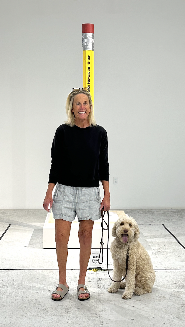 A woman with blonde hair and glasses on her head smiling while standing next to a fluffy dog in an art gallery. Behind them is a sculpture resembling a large pencil with a red eraser on top, a yellow body, and a silver tip, with the words "LIFE DEMAN