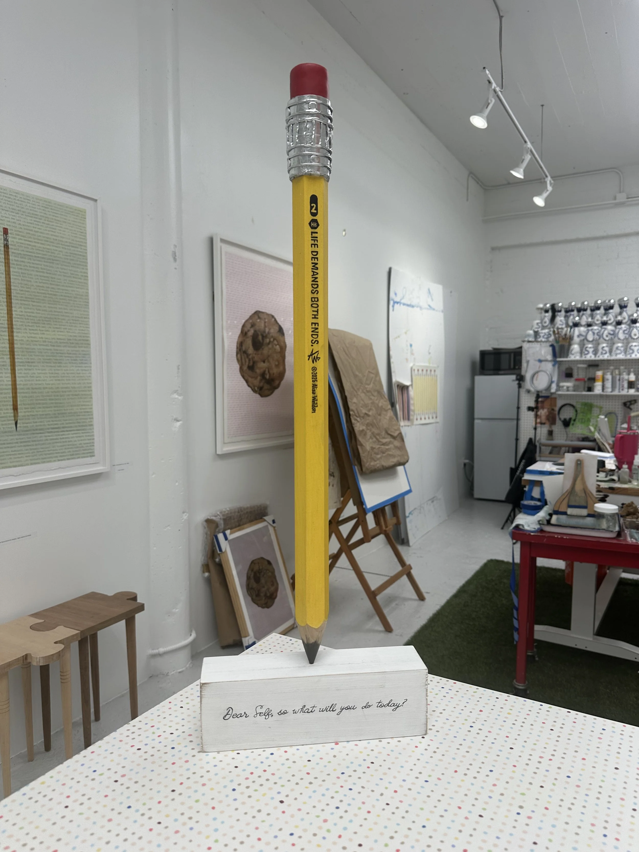 A large yellow pencil with a red eraser and metal ferrule balanced vertically on a white wooden block with the handwritten message, 'Dear Self, so what will you do today?'. The background shows art supplies and paintings in an art studio.