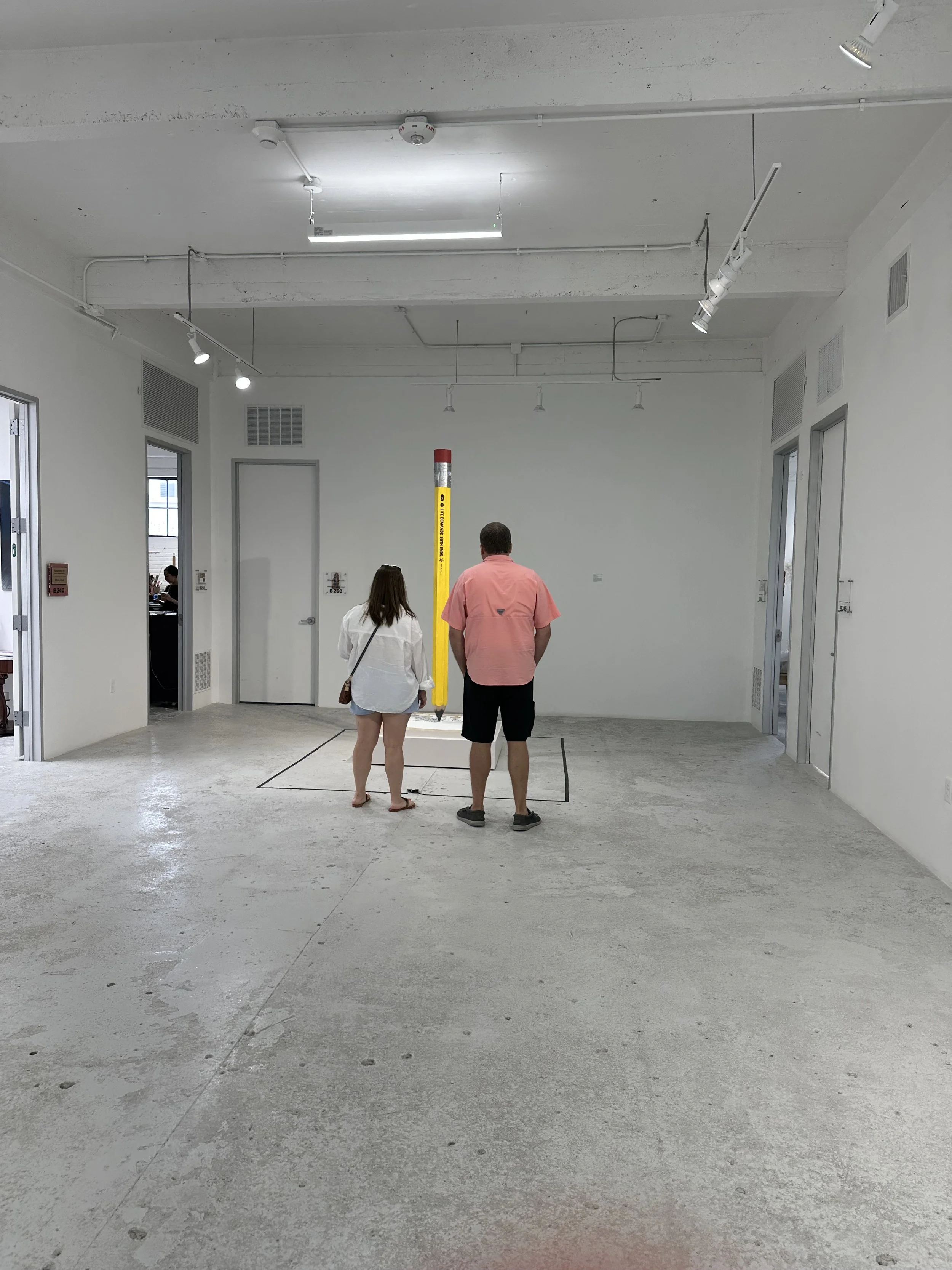 Two people, a woman and a man, standing indoors in front of a yellow safety pole in a room with white walls and ceiling, concrete floor, in a building that appears to be under construction or renovation.