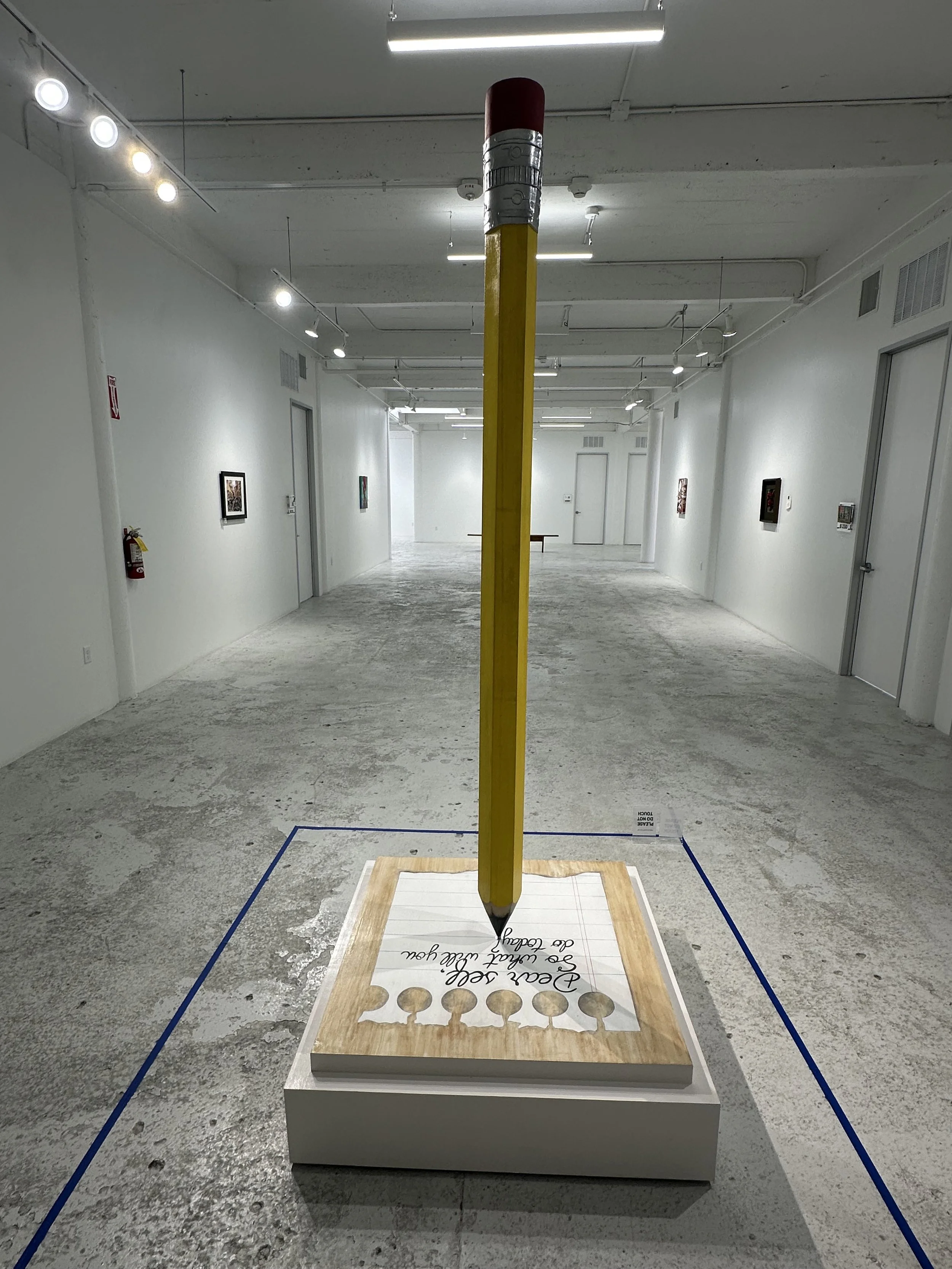 An art installation in a gallery featuring a large yellow pencil with a red eraser, embedded in a piece of wood with handwritten text, in a white-walled room with gallery lighting.