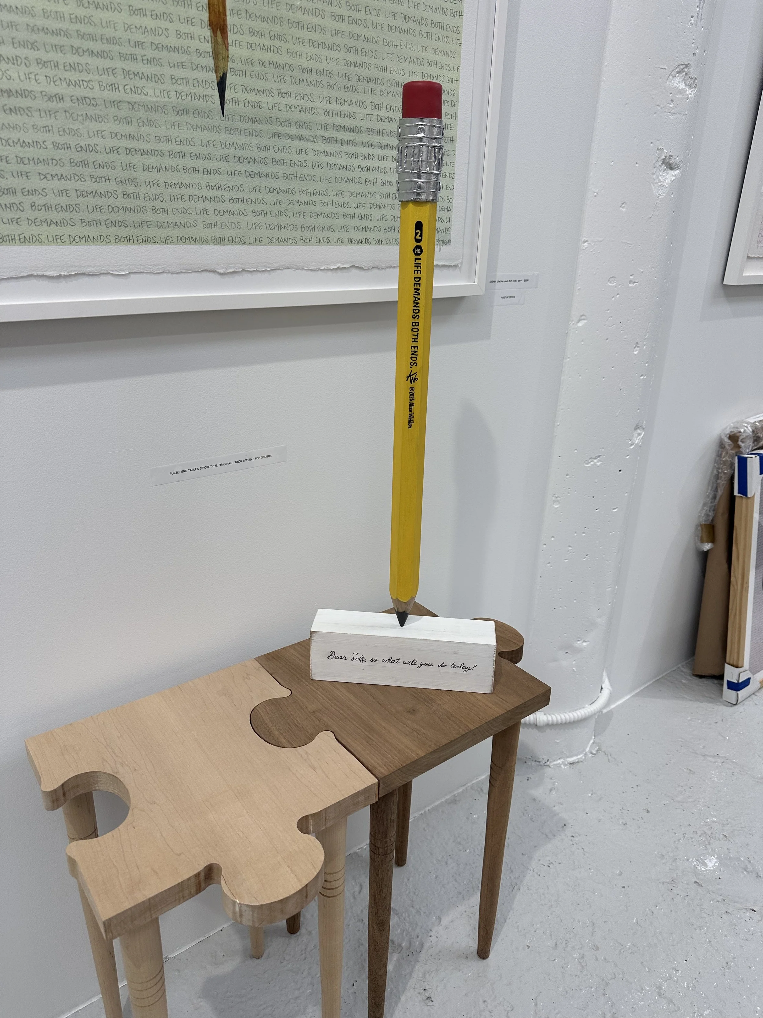 An art installation featuring a large yellow pencil with a pink eraser, standing upright on a small wooden table. The pencil has text printed on it reading "JULE DEMANDS BOTH ENDS!" and a small black and white logo. There is a block beneath the penci