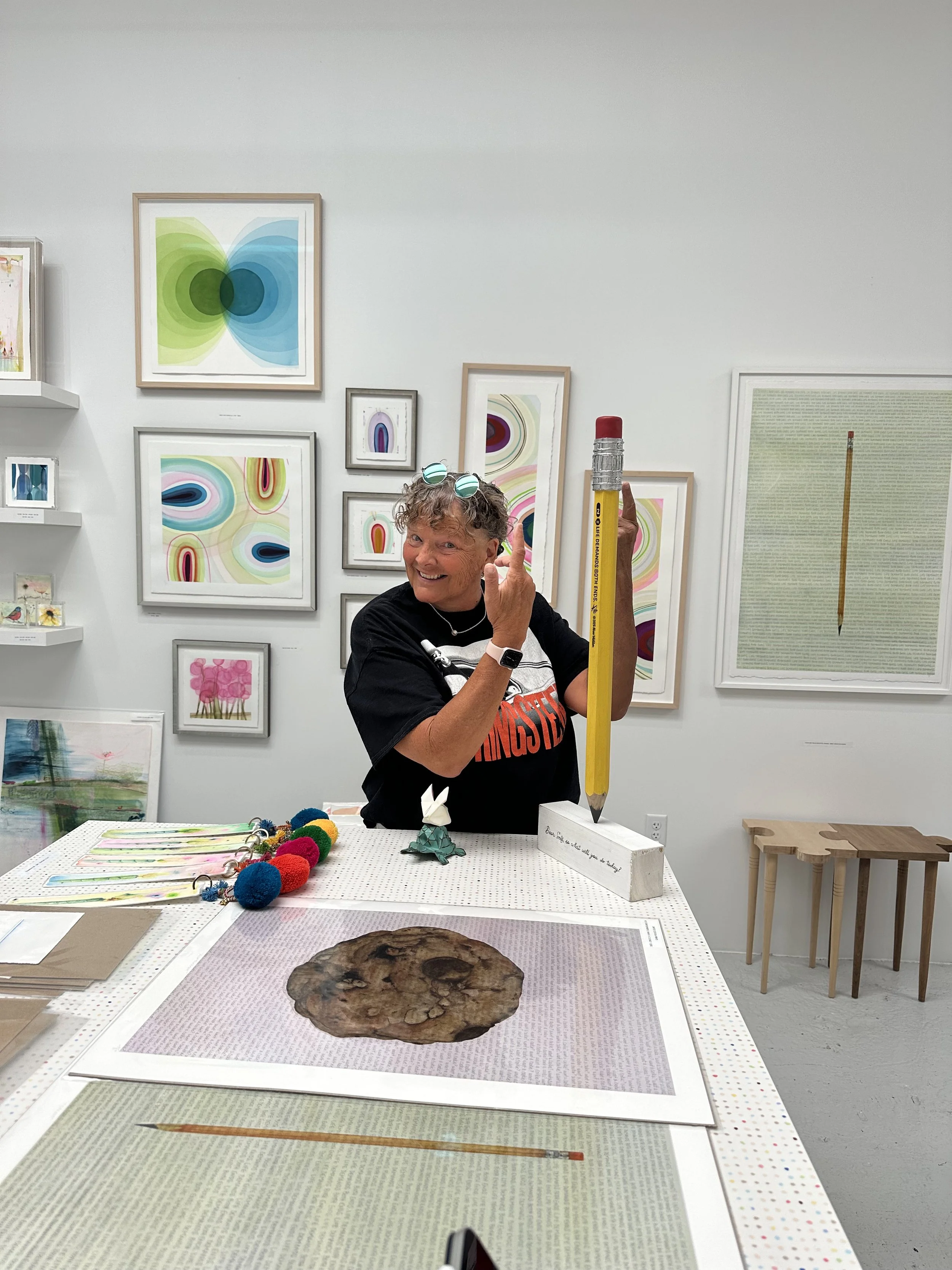 A woman standing at a table in an art gallery, holding a large yellow pencil with a red eraser tip, smiling and touching her face, surrounded by colorful abstract framed artworks on the wall.