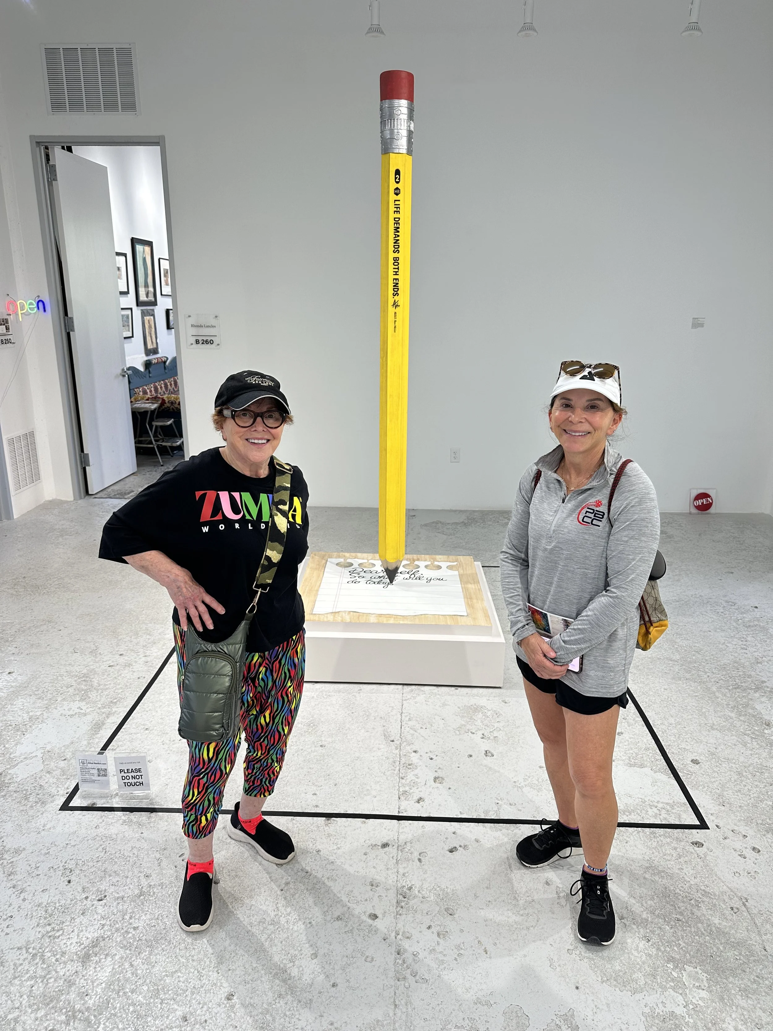 Two women standing next to a large yellow pencil sculpture with a batting and writing surface at the base, in an art gallery.