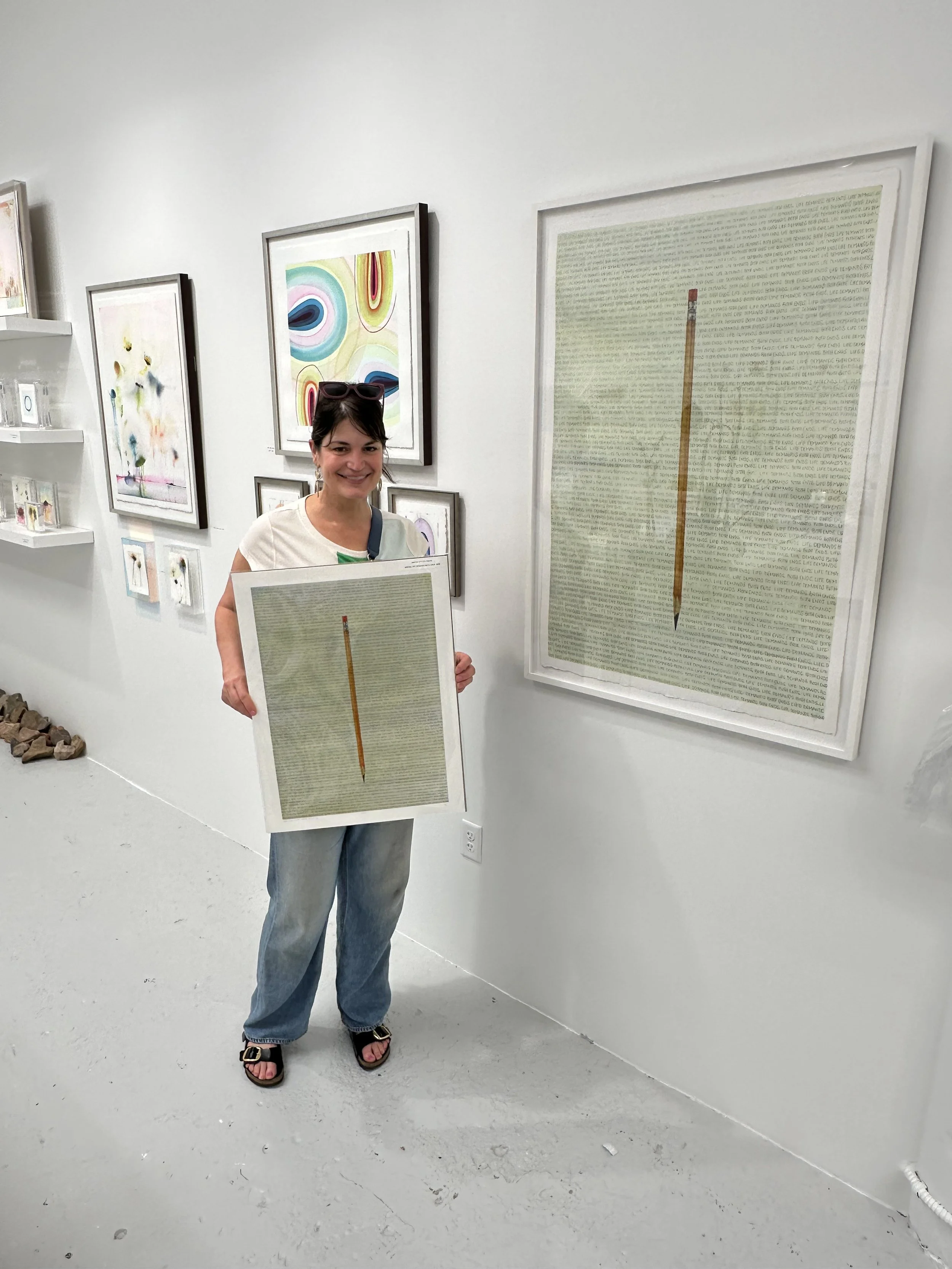 Woman with dark hair, sunglasses on her head, smiling, holding a framed art print of a pencil on a text-heavy background, inside an art gallery with abstract and colorful paintings on the wall.