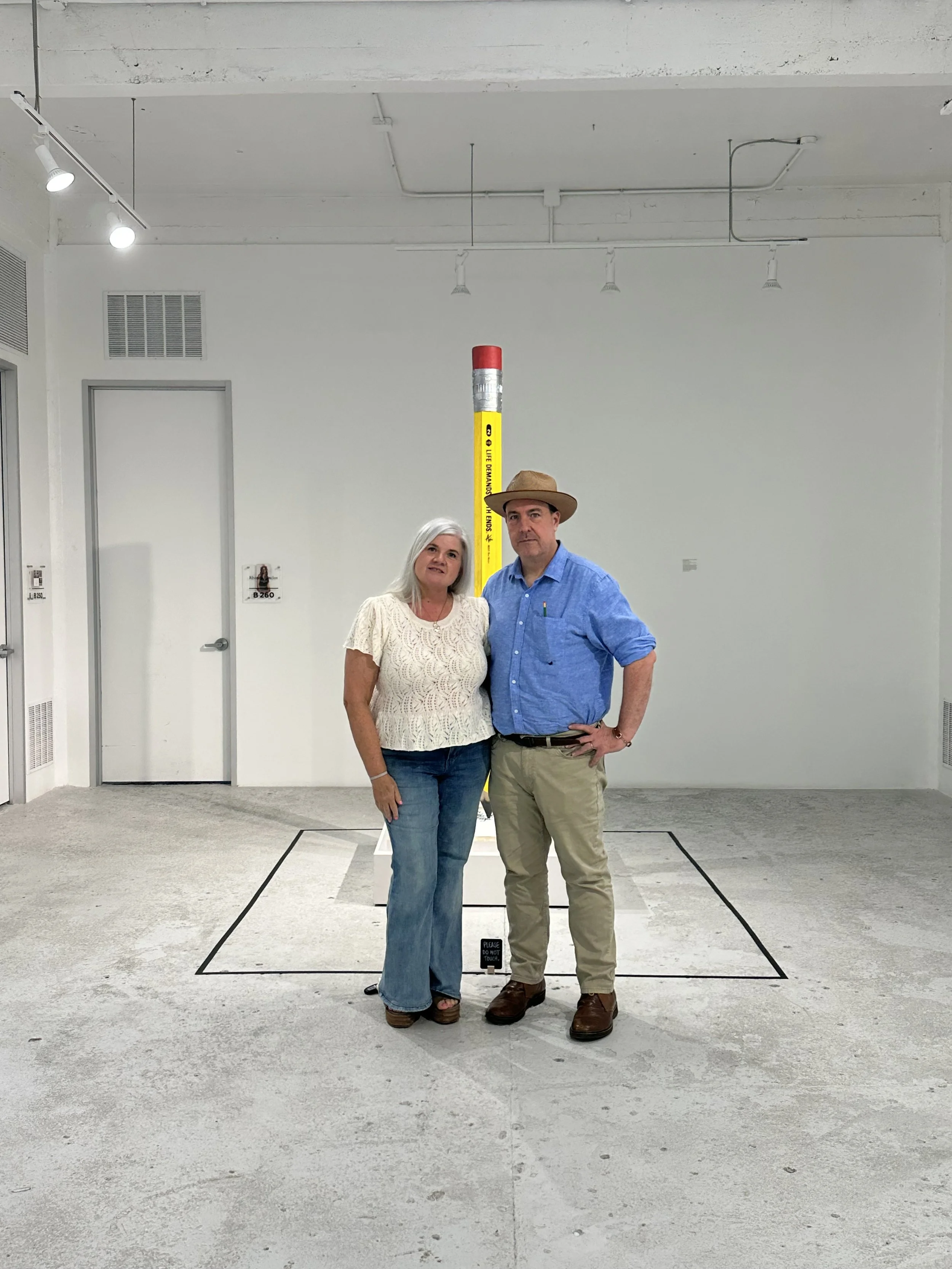 Two people standing in front of an art installation with a yellow pole and red top in a gallery space.