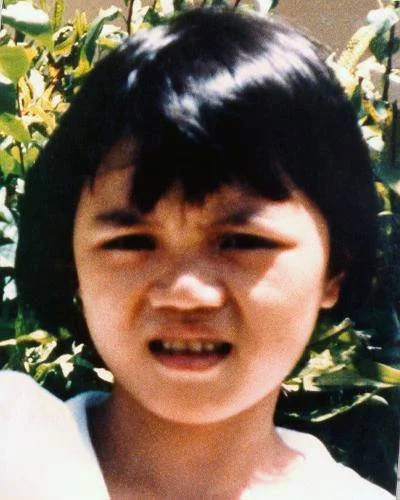 Childhood photo of missing girl, Jie Zhan Li, age unknown, wearing a white top with short black hair, brown eyes, standing outdoors in front of green foliage - vital reference image for long-term missing persons case, used in public awareness and ID