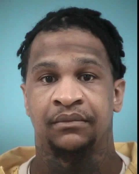 Mugshot of African American male with short twisted hair wearing an orange prison-issued garment, posed against a solid light blue background—arrested in connection with a murder investigation.