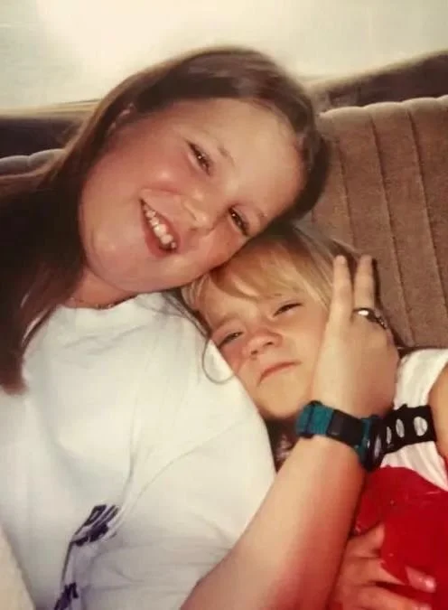 Close-up of a person wearing a white shirt and blue-strap wristwatch, seated on a brown couch with a silver ring on their finger—stylish minimalist fashion with subtle red accent in corner.