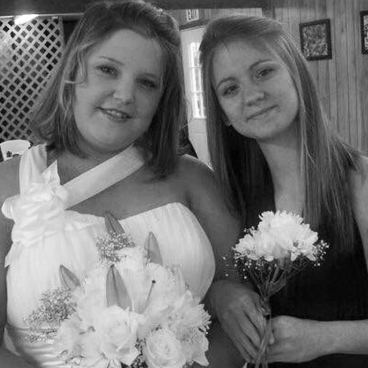 Two individuals standing close indoors, holding floral bouquets—one in a white wedding dress with shoulder embellishment, the other in a dark formal gown—framed by rustic wooden walls and vintage decor, evoking intimate wedding or celebration vibes.