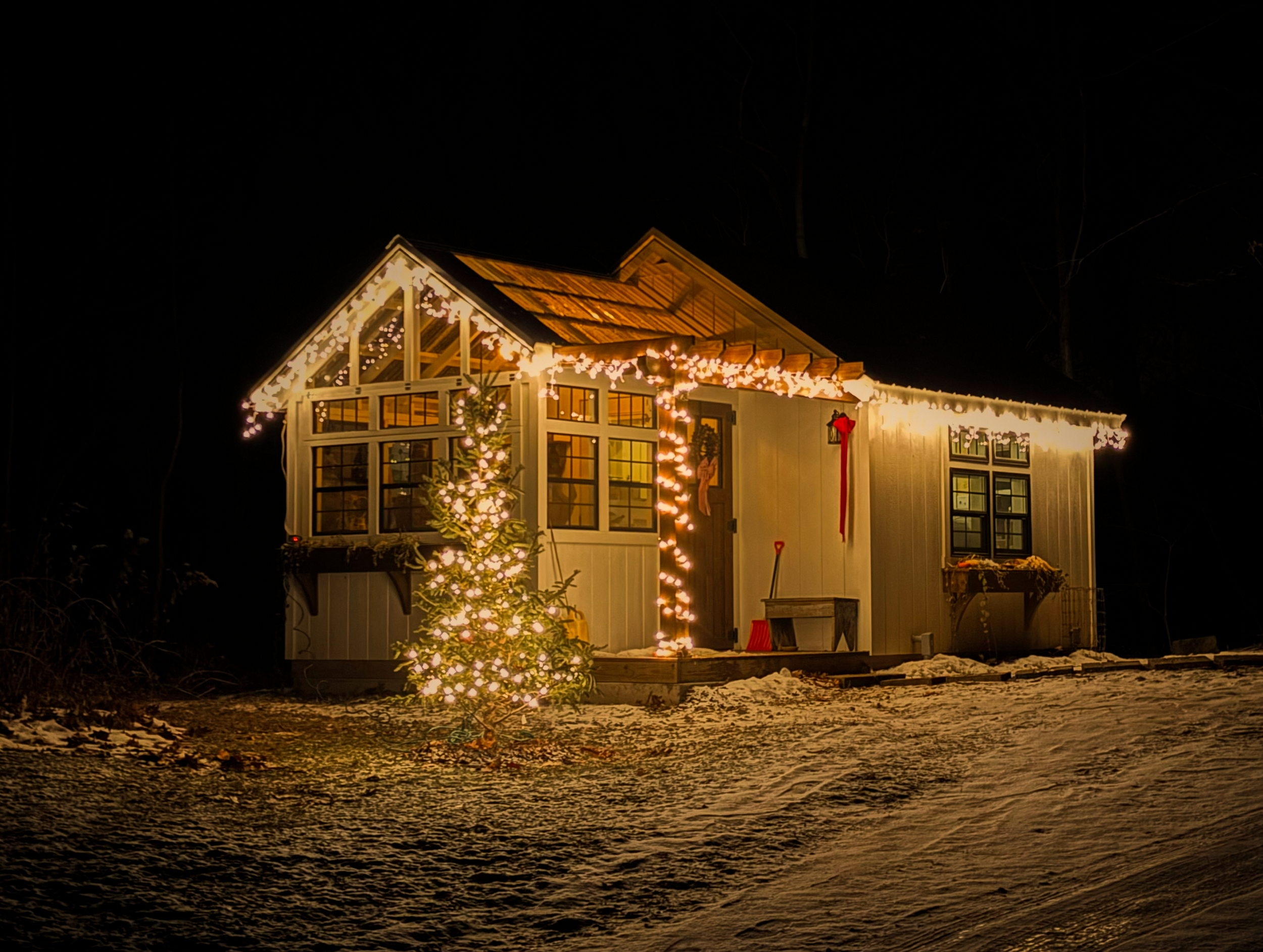 A warmly lit home in winter with Christmas lights outlining the roof, a glowing tree in the yard, and fresh snow—capturing a nostalgic, storybook holiday evening.