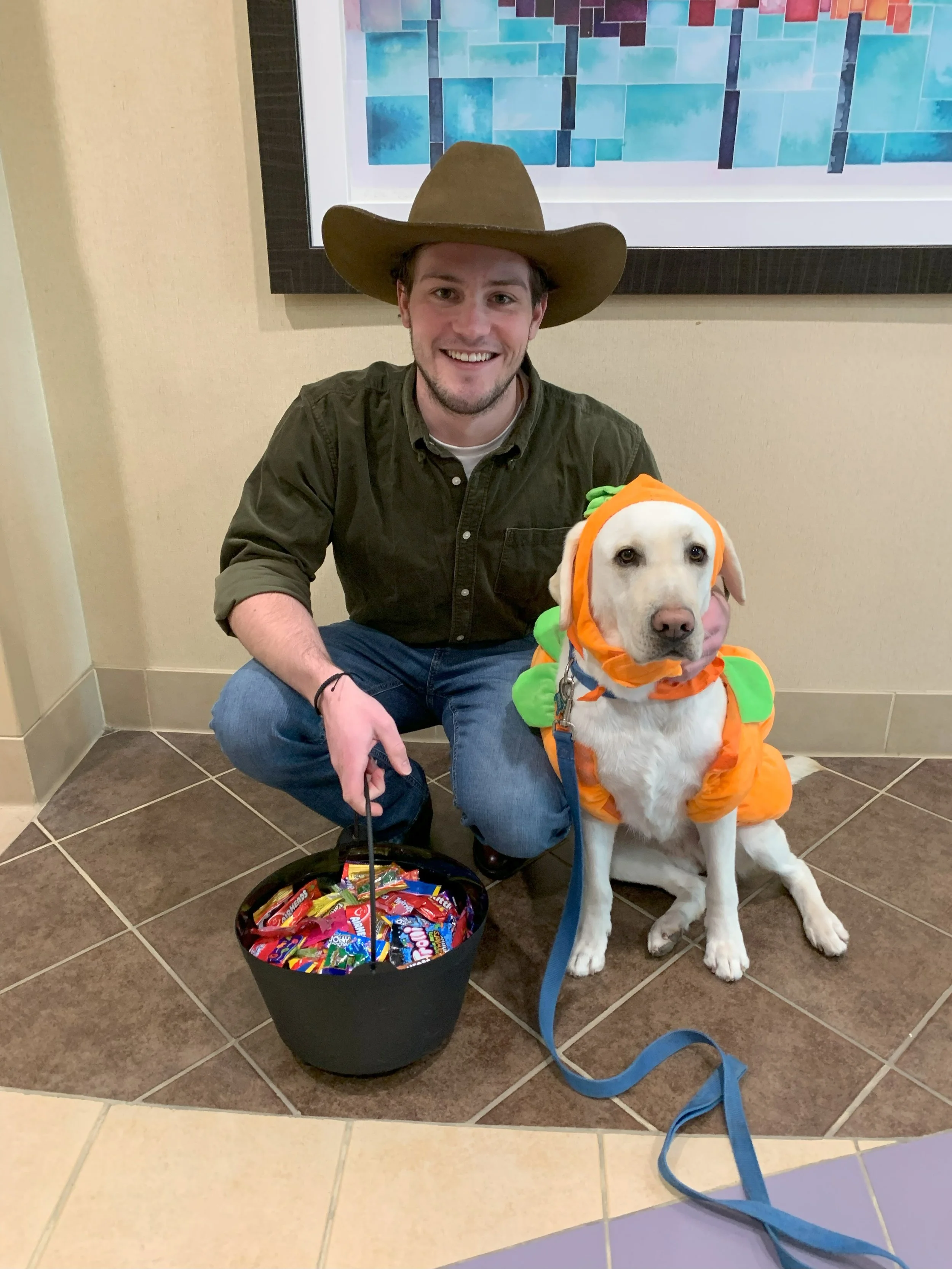 A man smiling, wearing a cowboy hat, kneeling next to a dog dressed in a pumpkin costume, with a basket of candy on the floor.