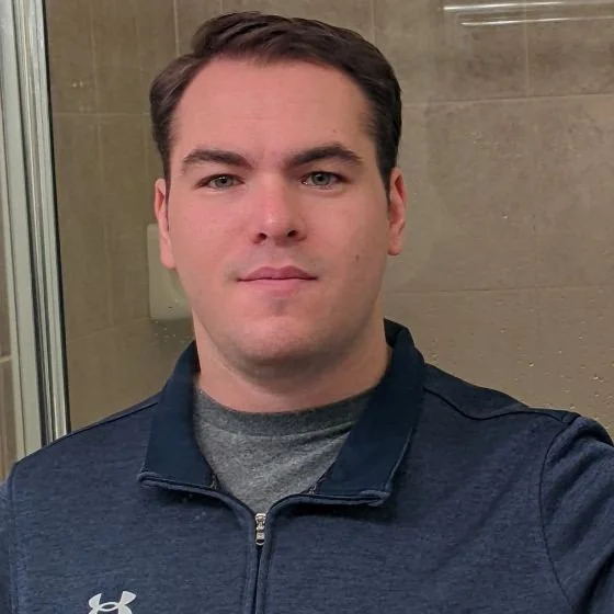 A young man with short dark hair and fair skin taking a selfie indoors, wearing a navy blue zip-up jacket over a gray shirt, with beige tiled wall background.
