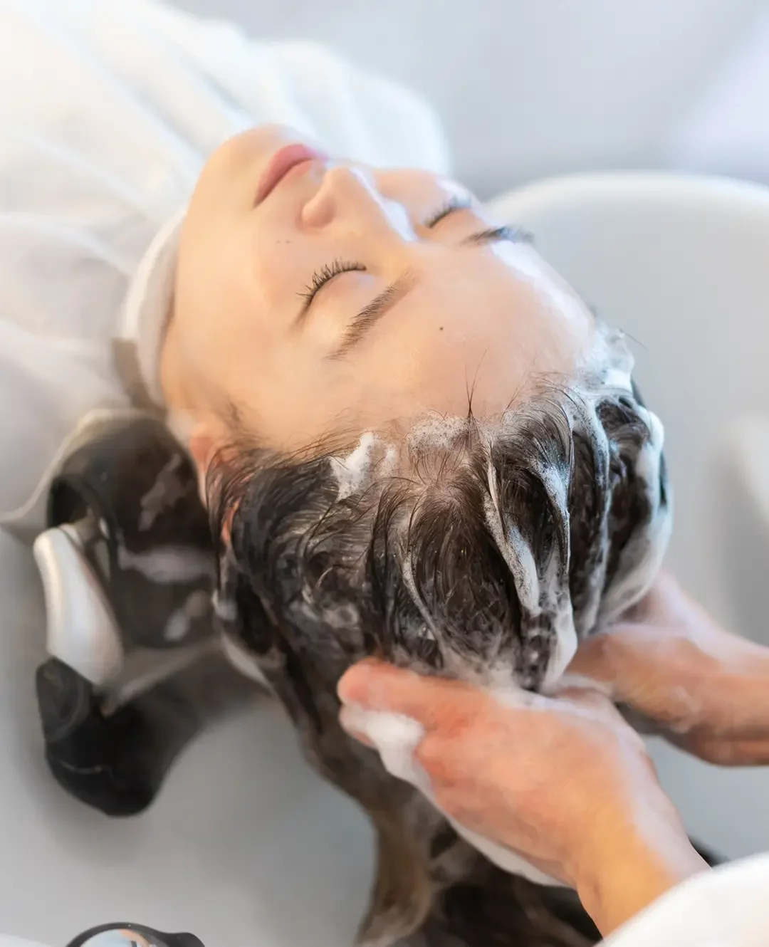 Woman with eyes closed at salon sink receiving a moisturizing, color-safe deep conditioning treatment from a stylist.