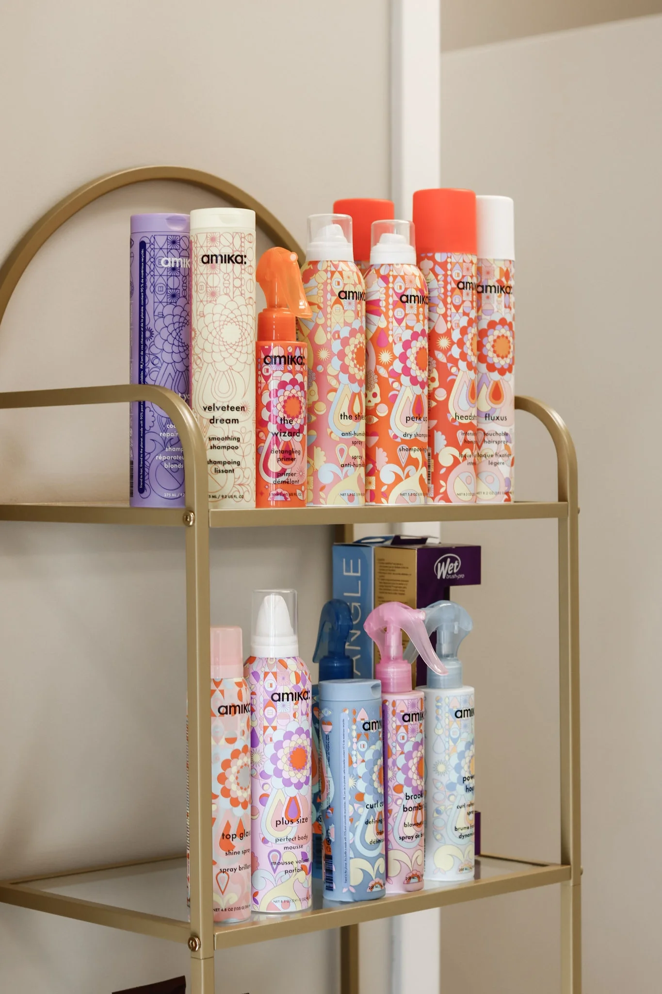 Amika Plus Size volumizing spray, The Wizard detangler, Top Gloss Shine Spray, Curl Corps Curl Defining Cream, Perk Up dry shampoo, and other volumizing products displayed on a gold shelf.