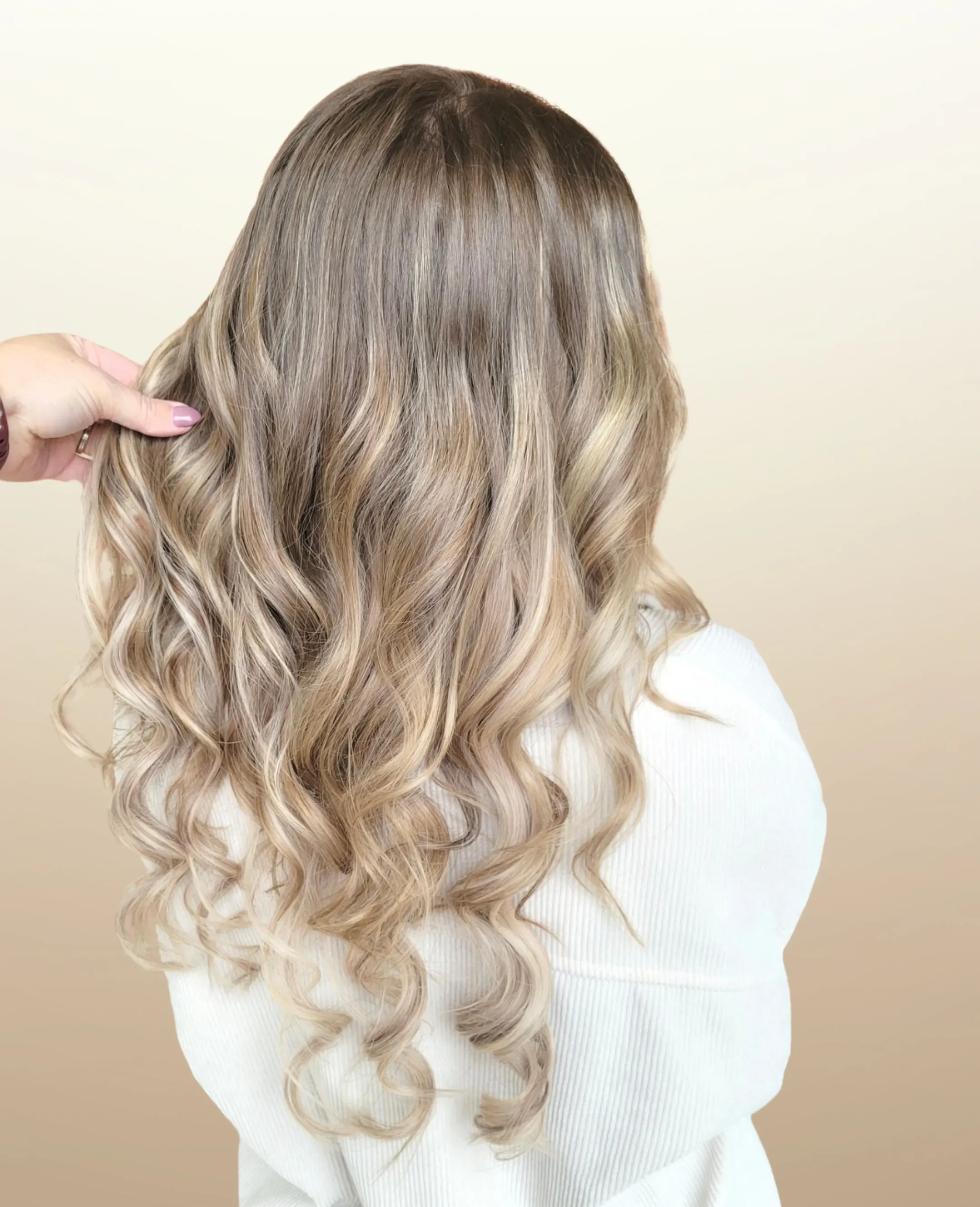 Hand holding a section of long blonde hair with loose mid-length curls and seamless extensions.