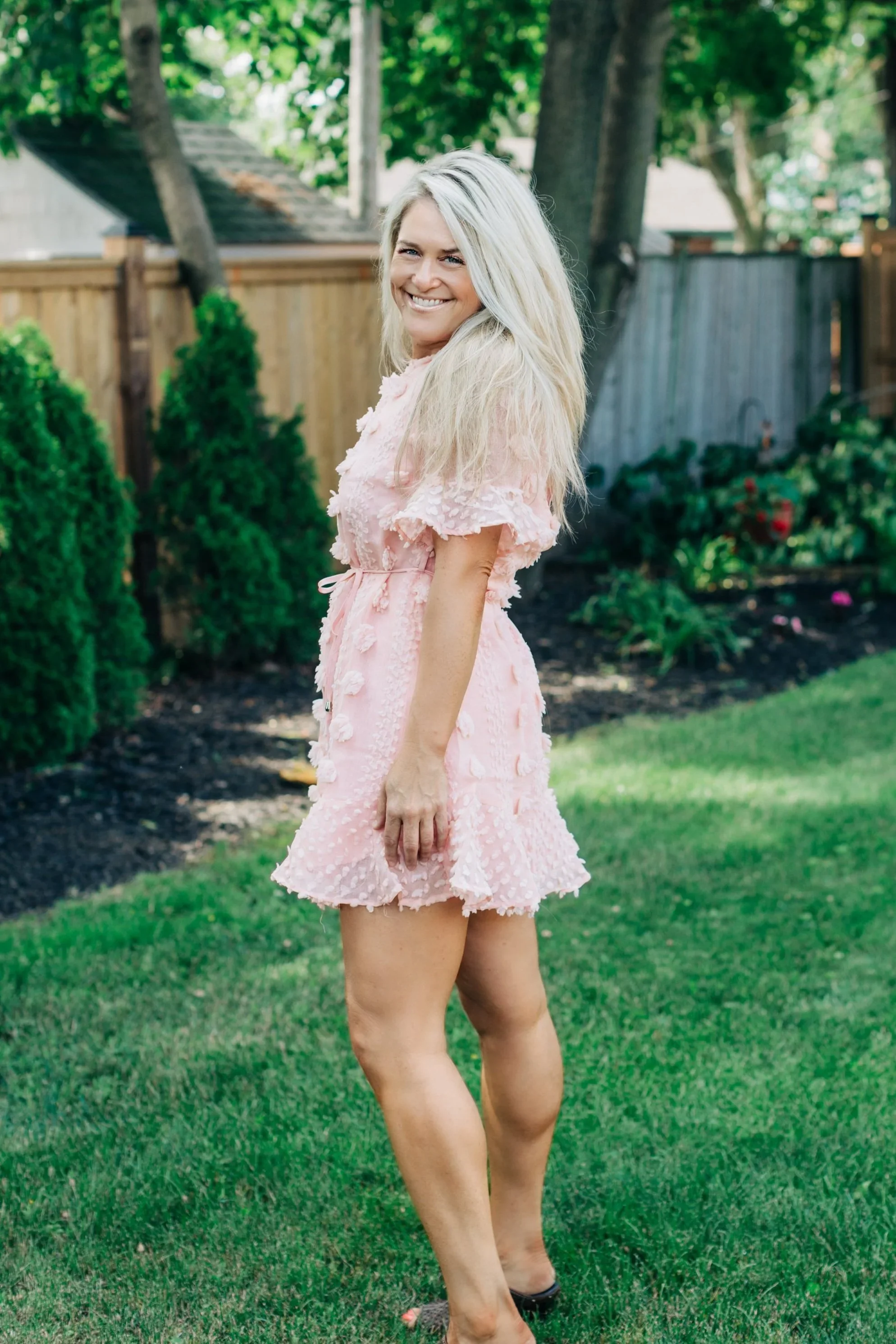 A woman with long blond hair smiling and standing in a backyard garden, wearing a pink dress with textured floral details, surrounded by greenery and trees.