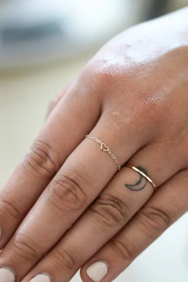 Close-up of a person's hand showing three rings and a small tattoo of a crescent moon on the finger. The hand has painted nails and the background is blurry.