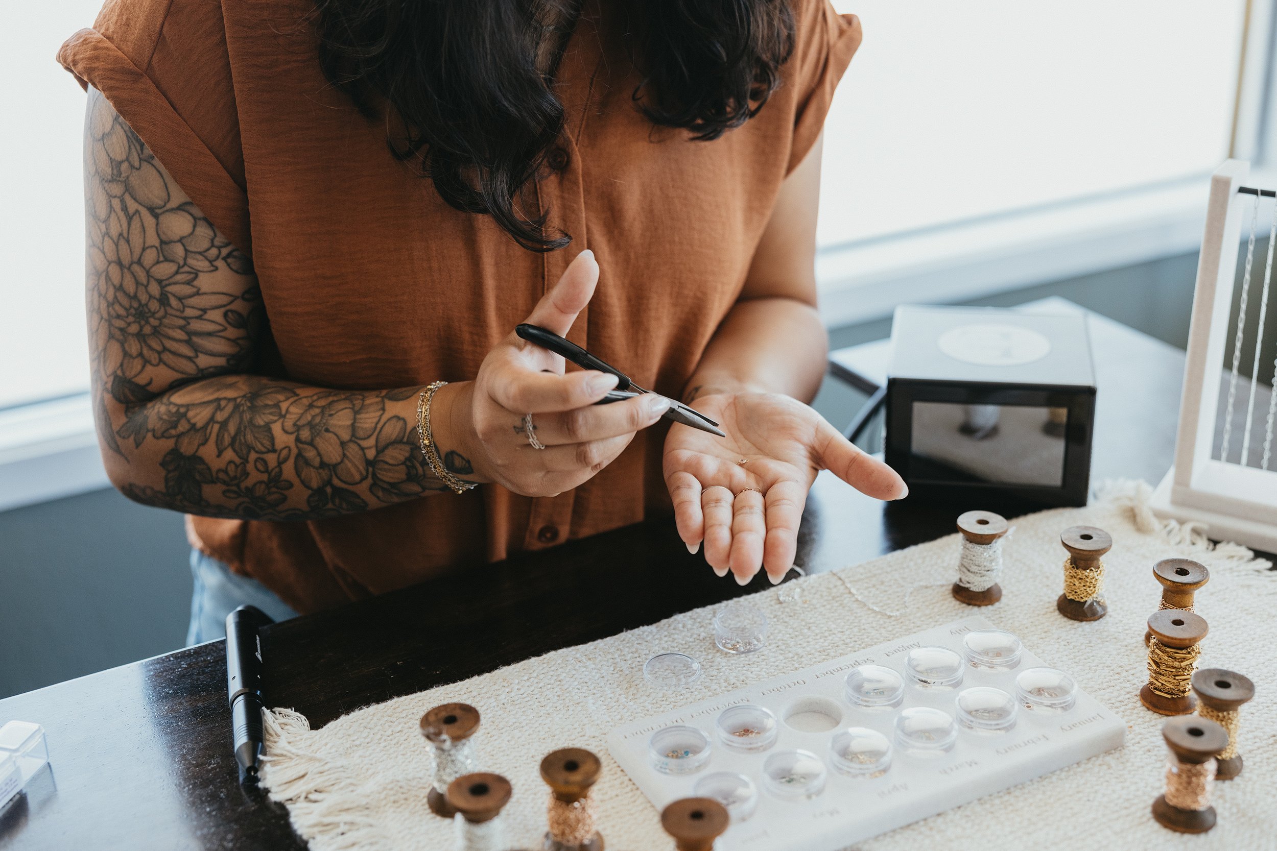 Woman with tattooed arms making jewelry, holding a small item in her left hand and using a tool in her right hand on a worktable with spools of thread and other jewelry-making supplies.