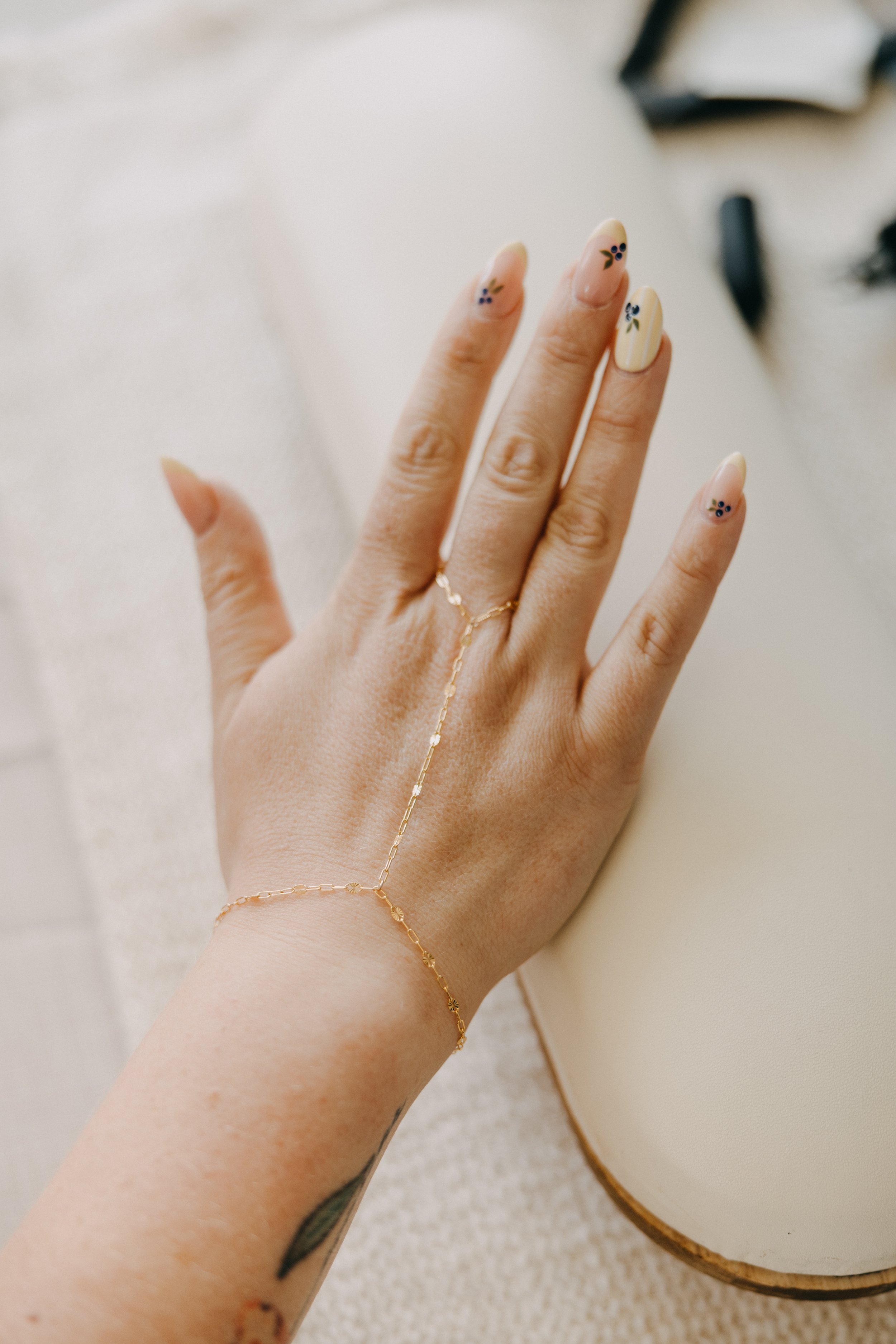 Close-up of a hand with a delicate chain bracelet that drapes to form a T shape, with a textured fabric in the background.