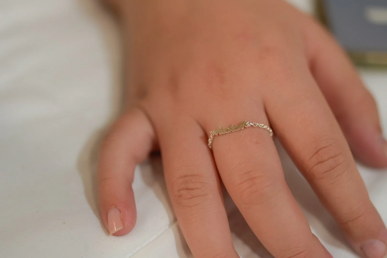 Close-up of a person's hand showing three rings and a small tattoo of a crescent moon on the finger. The hand has painted nails and the background is blurry.