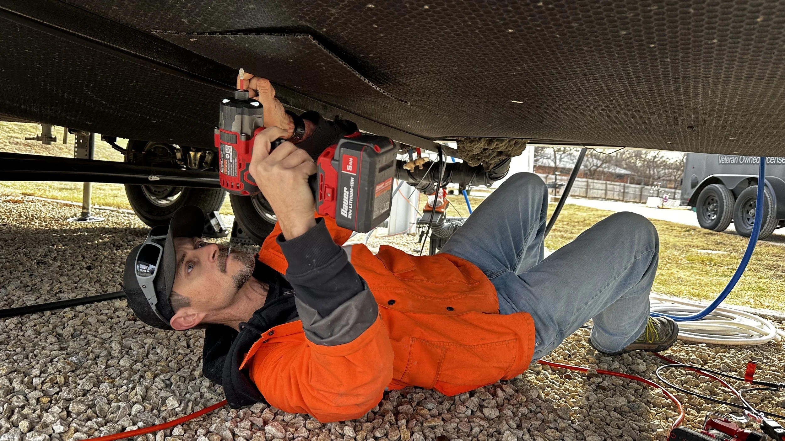 Mobile RV Tech lying on gravel ground working underneath an RV, using a power drill, wearing an orange and black jacket, gray pants, and a black cap.