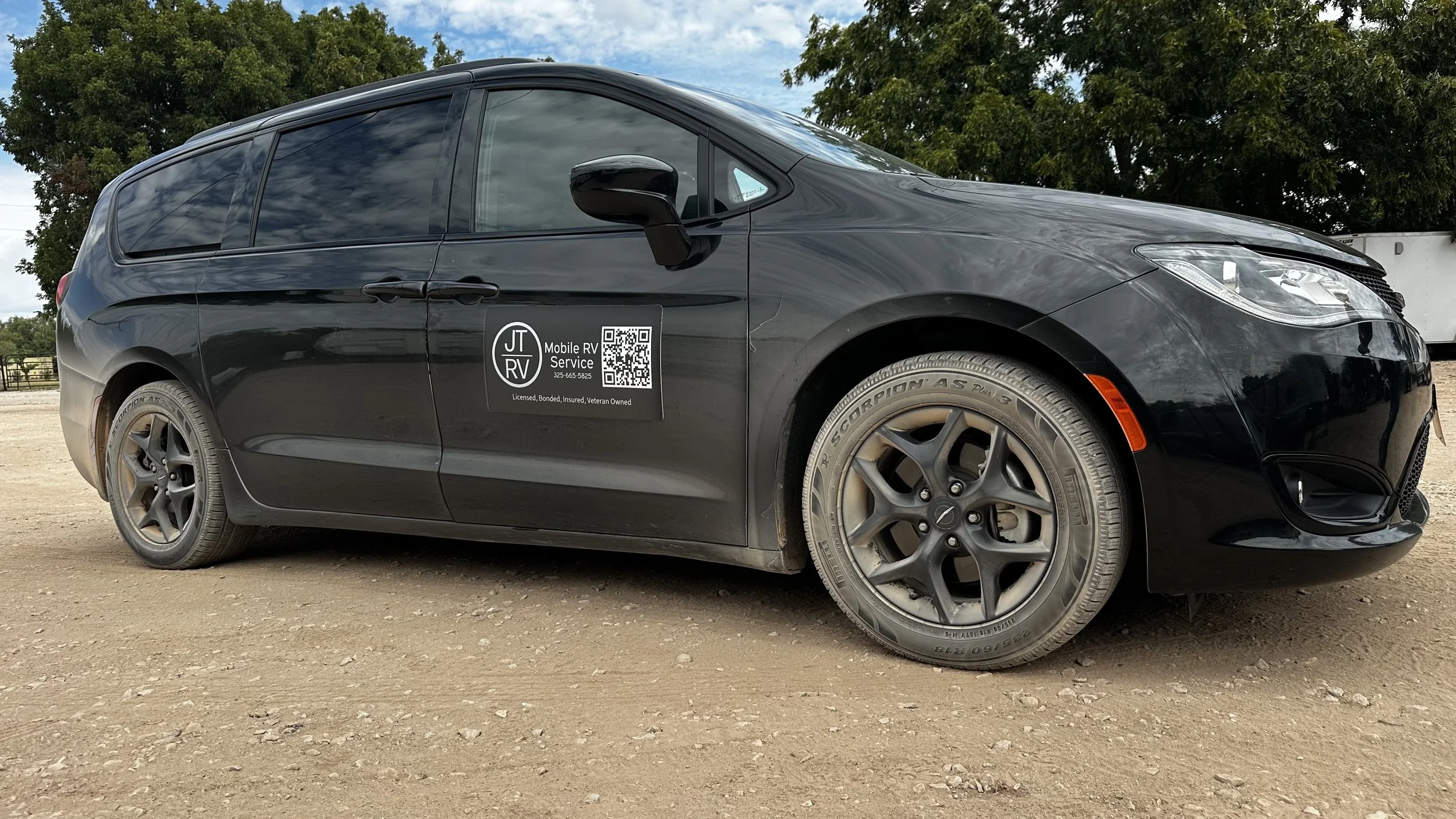 Black mobile RV tech minivan  with advertising sign on the door. The sign reads "JT RV Mobile RV Service" with a QR code and contact information. The vehicle is parked on dirt ground, with green trees and a fence visible in the background.