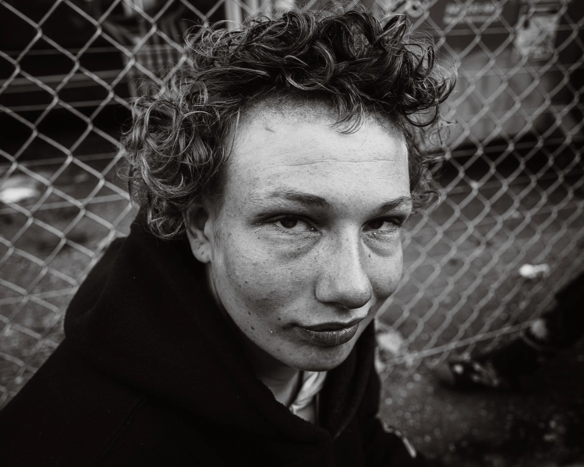 Close-up of a woman's face with curly hair, looking at the camera, with a chain-link fence in the background, in black and white.