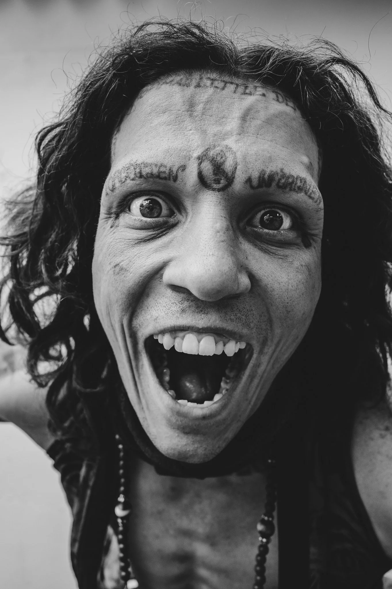 Black and white photo of a woman's face with tattoos on her forehead, wide-open mouth, and expressive eyes. She has curly hair and is wearing a necklace.