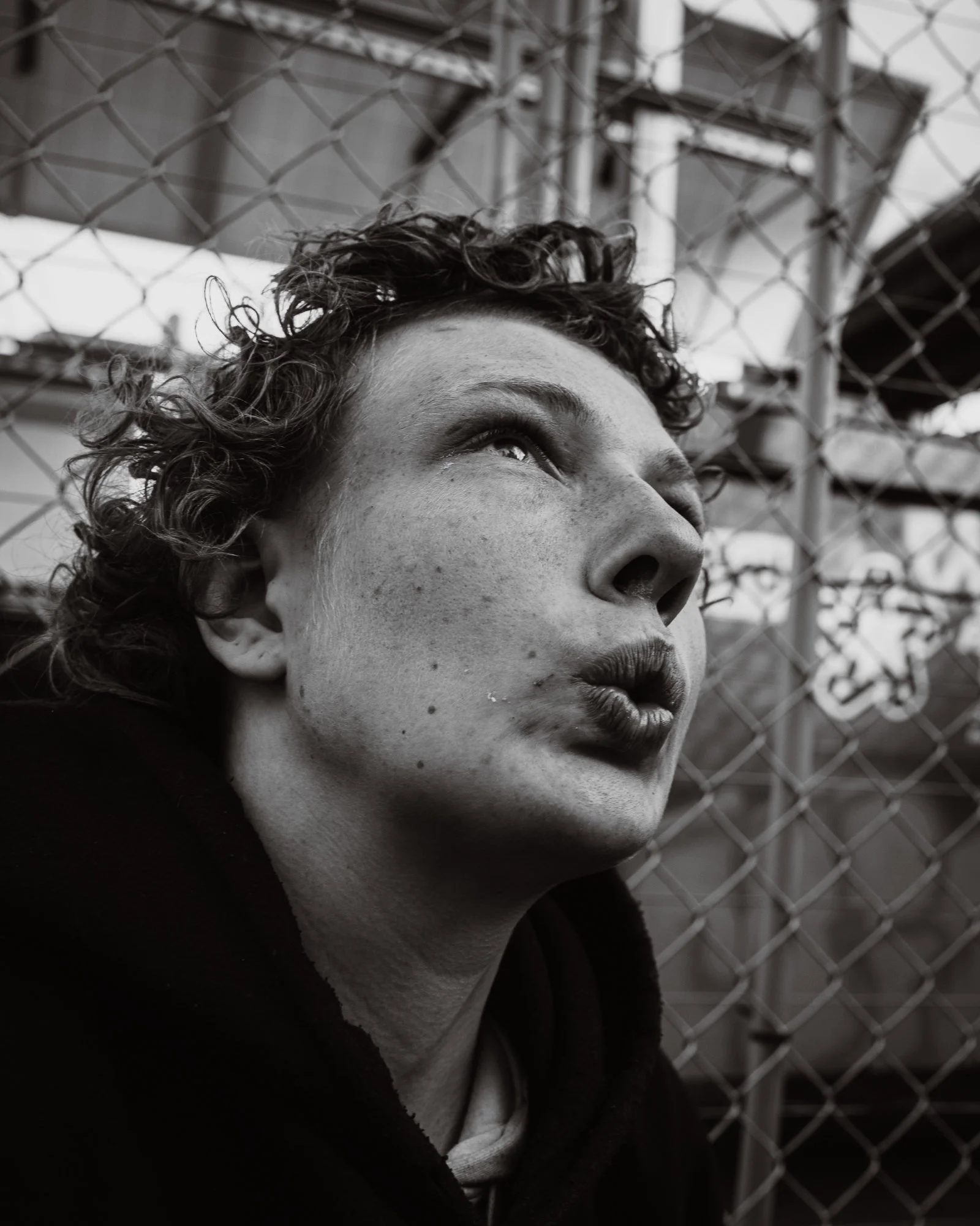 A young woman with curly hair and freckles looking upward, standing in front of a chain-link fence.