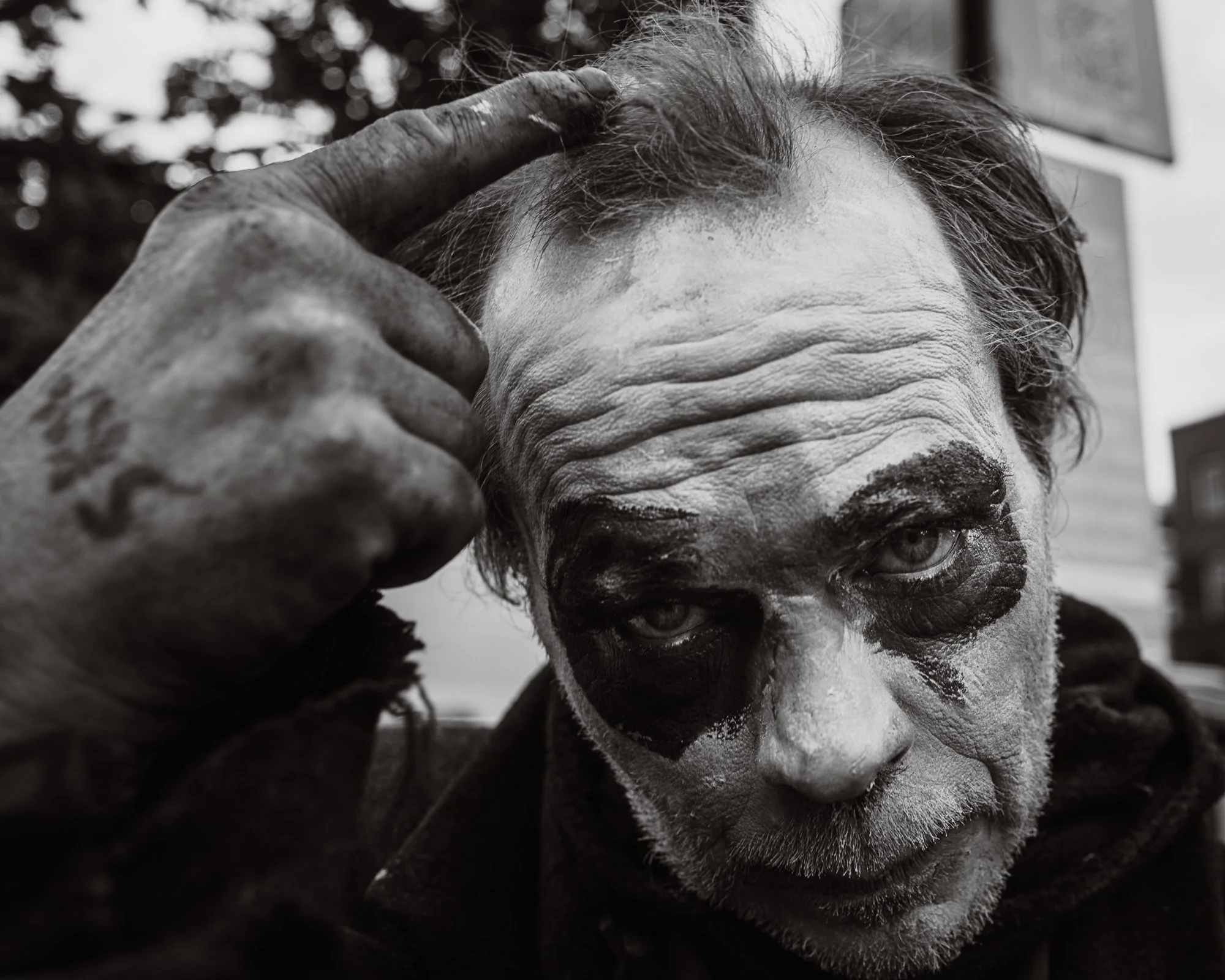 Close-up of a person with a painted face, possibly a clown or performer, making a gesture with fingers near the forehead. The person has curly hair and is looking downward or to the side.