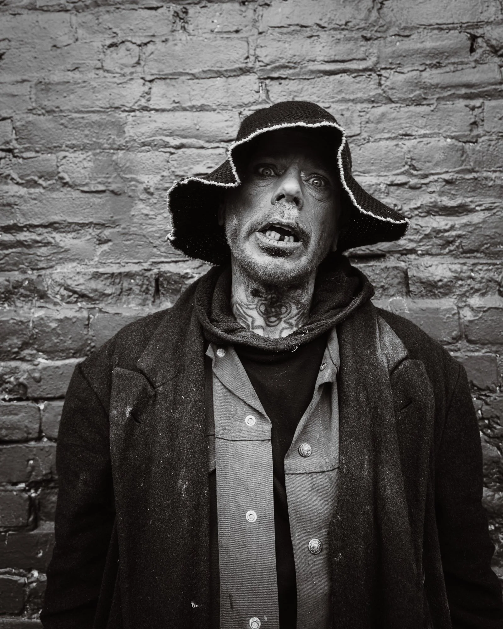 A black and white photo of a man with rugged features, hollow cheeks, and patchy facial hair, wearing a hat and a coat, standing in front of a brick wall.