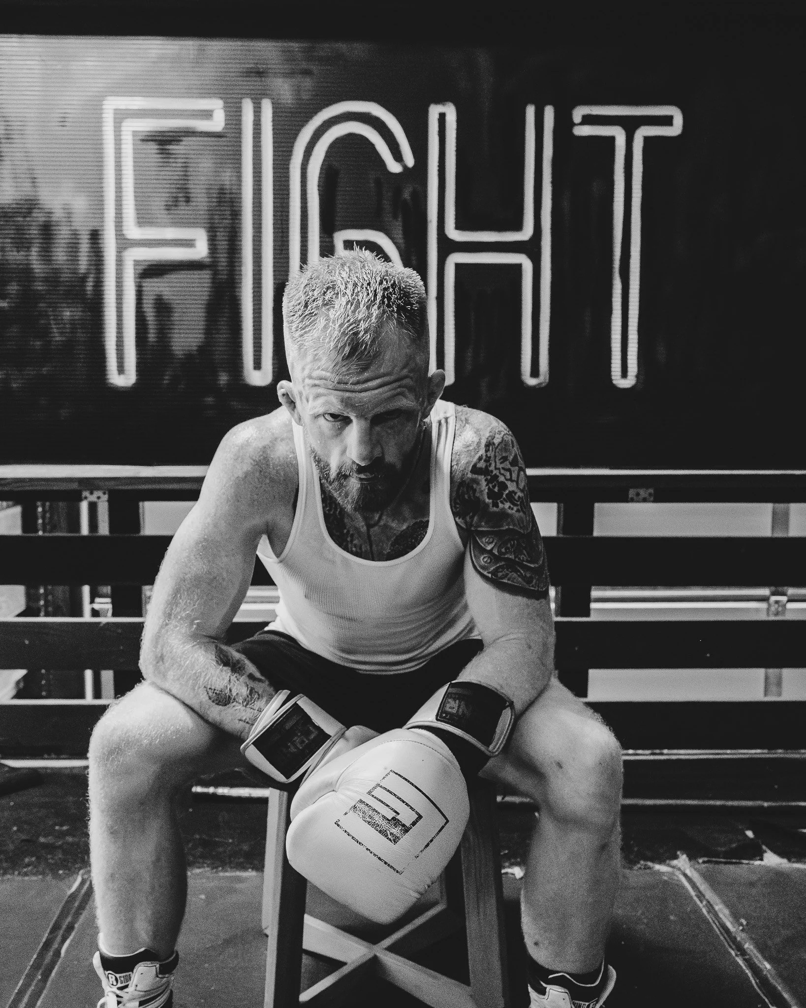 A muscular man with short hair and tattoos on his arm and chest sitting on a bench in a gym, looking into the camera, with a punching bag and foam roller behind him.