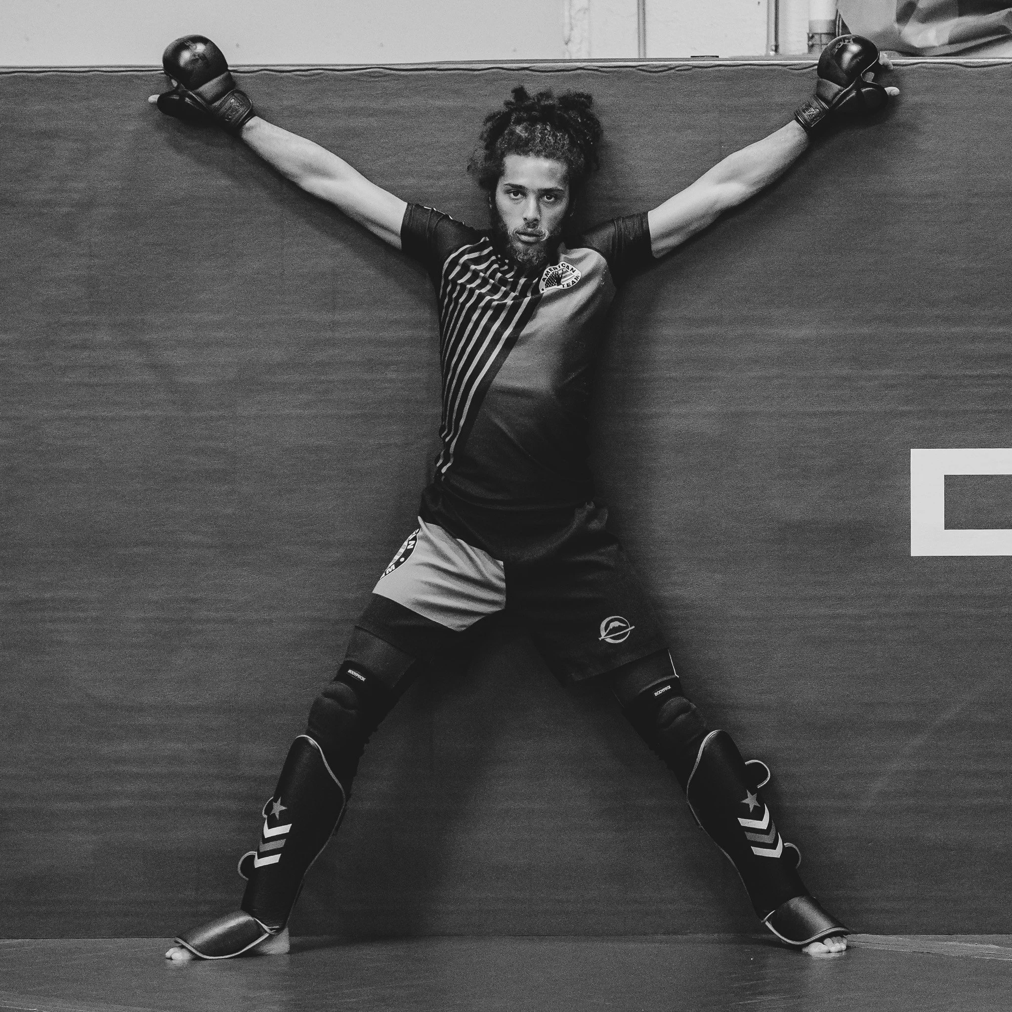 A person with curly hair and a beard posing with arms and legs spread out on a wall, wearing athletic clothing and gloves, in a black-and-white photo. MMA photography