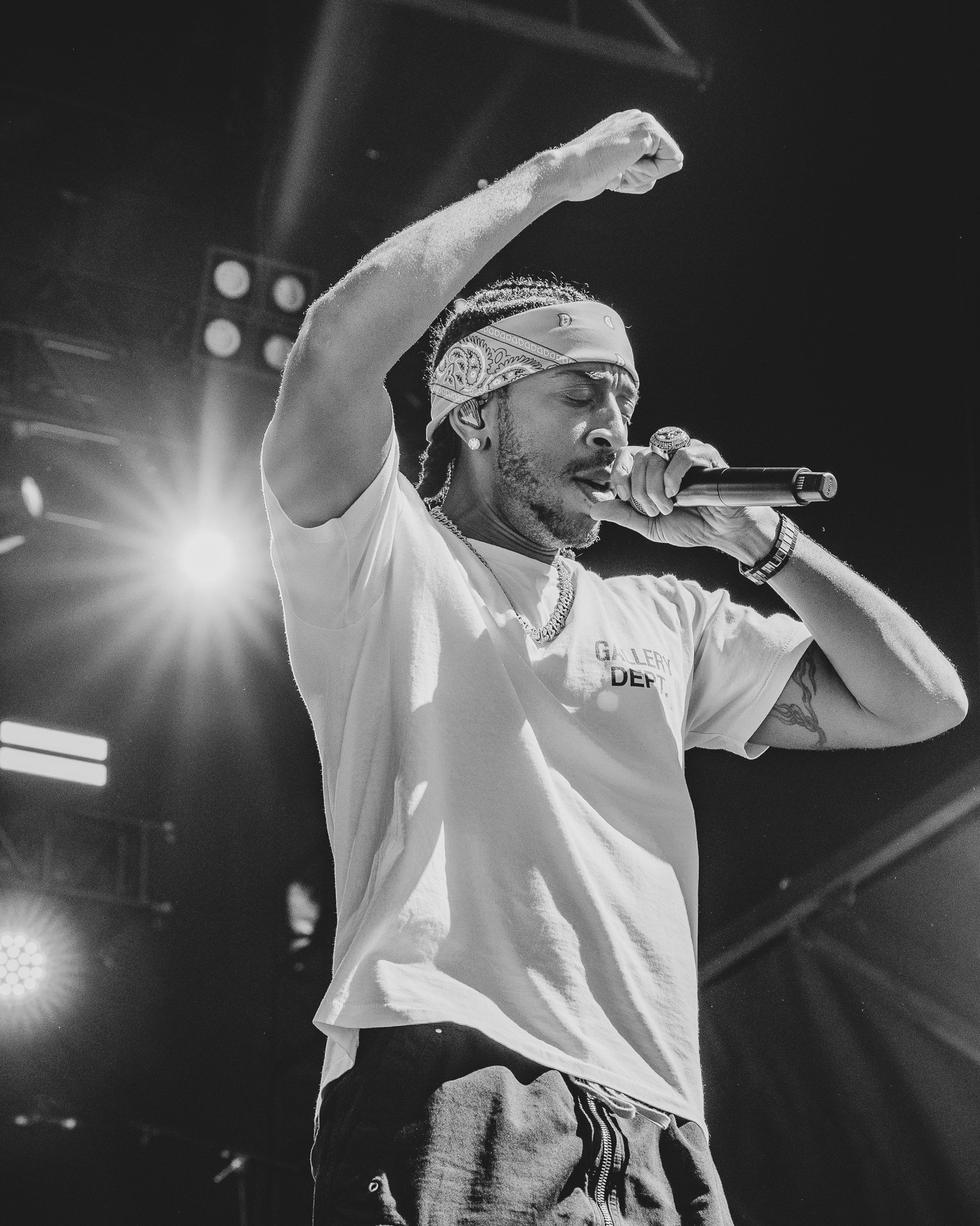 A man performs on stage, holding a microphone close to his mouth, with his eyes closed and his right arm raised. He wears a bandana, earrings, a T-shirt, and a watch. Stage lighting and equipment are visible in the background. Ludacris
