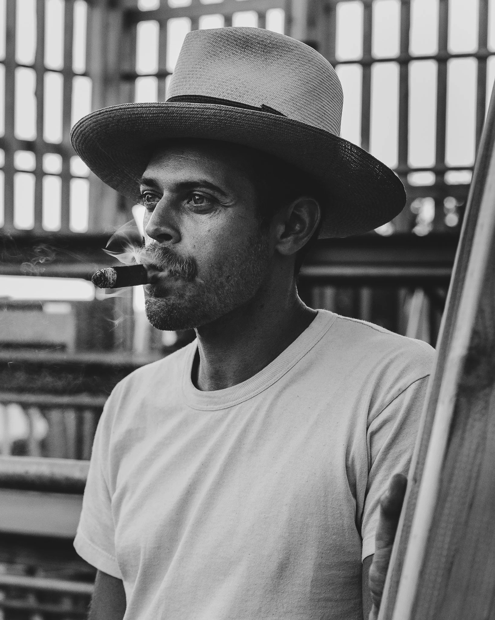 Man wearing a wide-brimmed hat and smoking a cigar, standing in front of a lattice fence.
