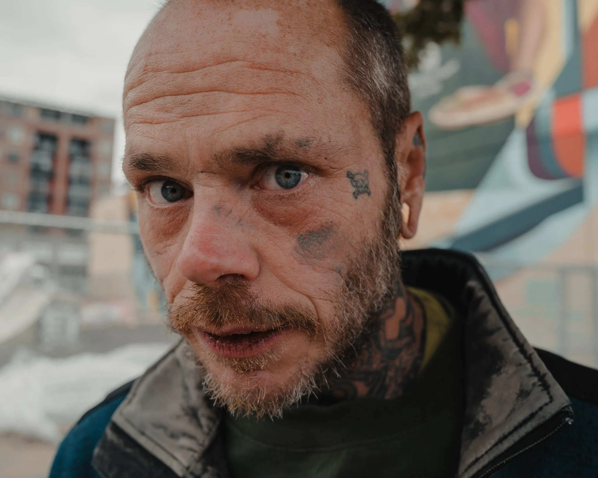 Close-up of a man with blue eyes, facial tattoos, and a beard, wearing a jacket, with buildings and colorful mural in the background.