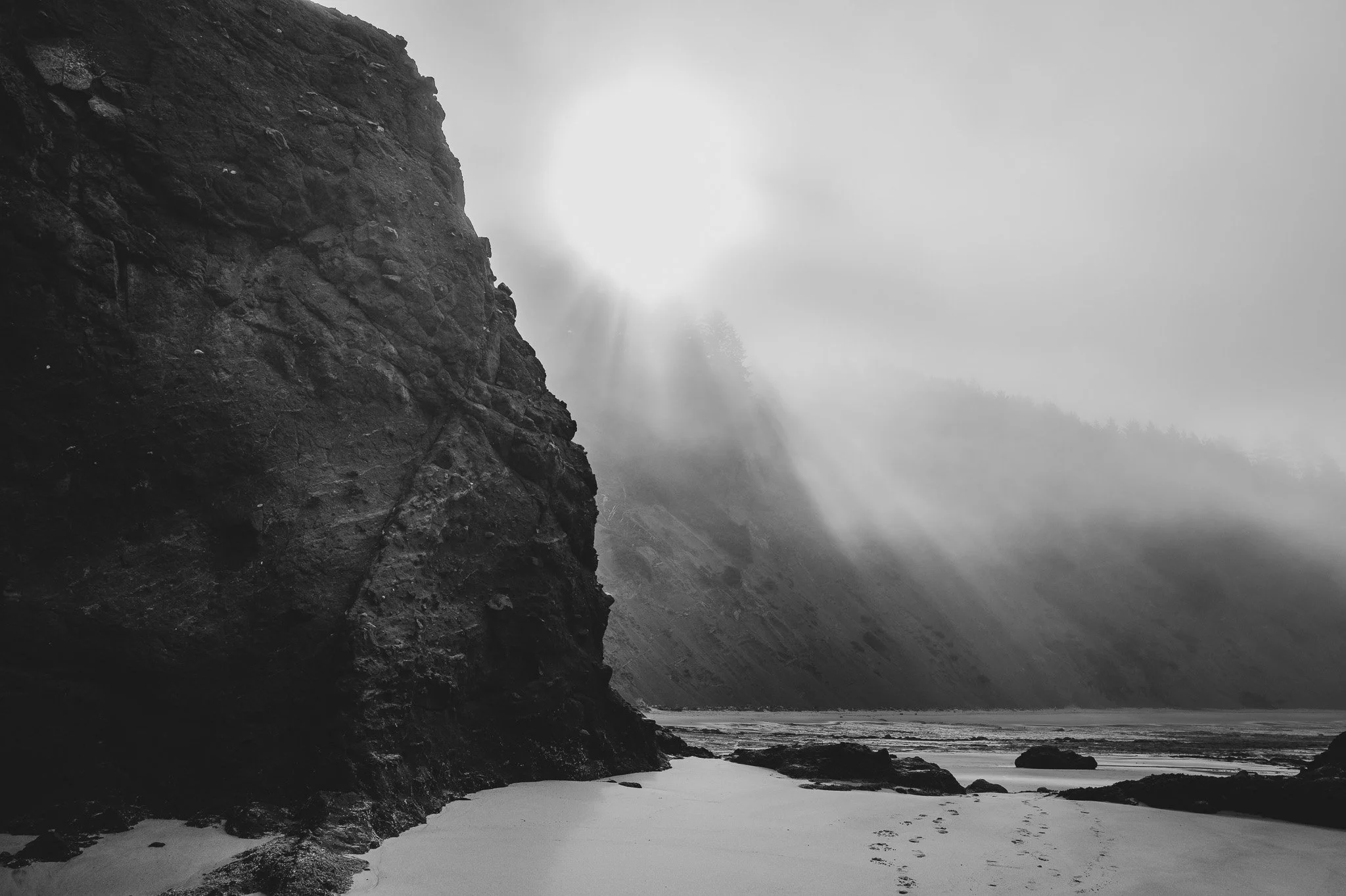 Black and white photograph of a rocky cliffside overlooking a foggy beach with large rocks and water in the distance.