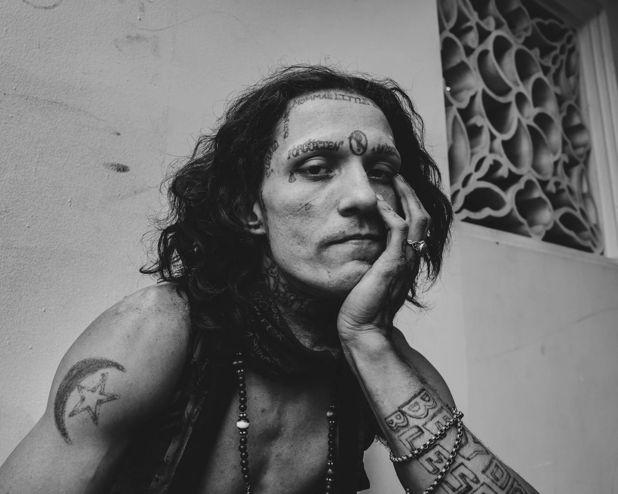 A black and white photo of a woman with tattoos on her face and arms, wearing jewelry and a bandana around her neck, sitting against a plain wall with patterned latticework in the background.