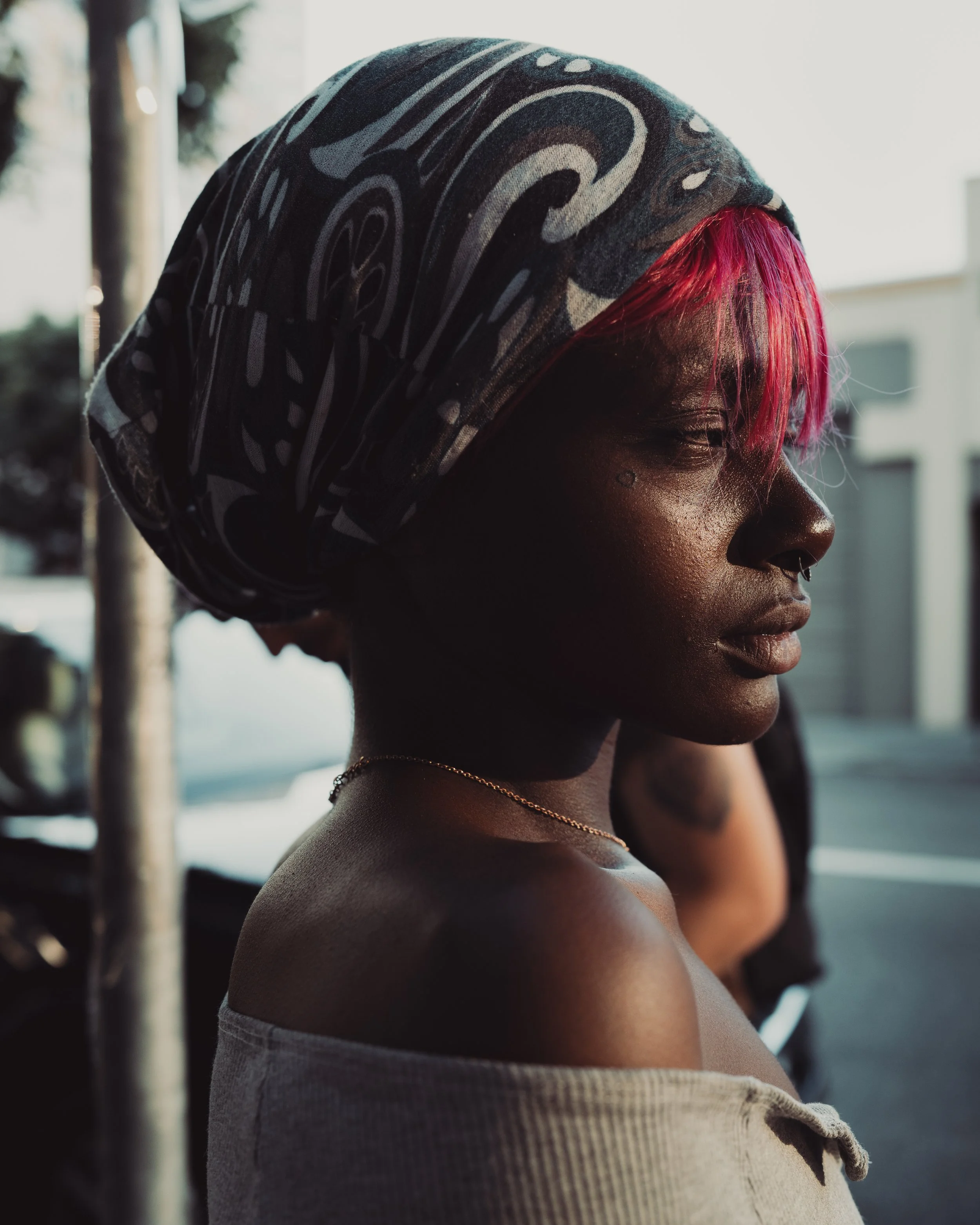 Profile of a woman with dark skin, bright pink hair, wearing a patterned headscarf, outdoors during sunset.