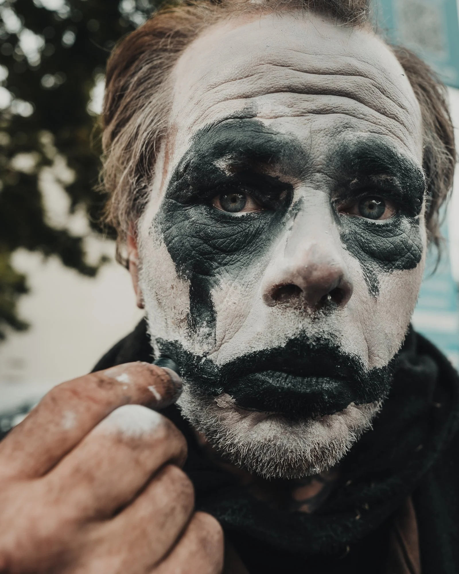 Close-up of a person's face with painted clown makeup, white face paint, black around the eyes, and black lips, holding a finger near the chin.