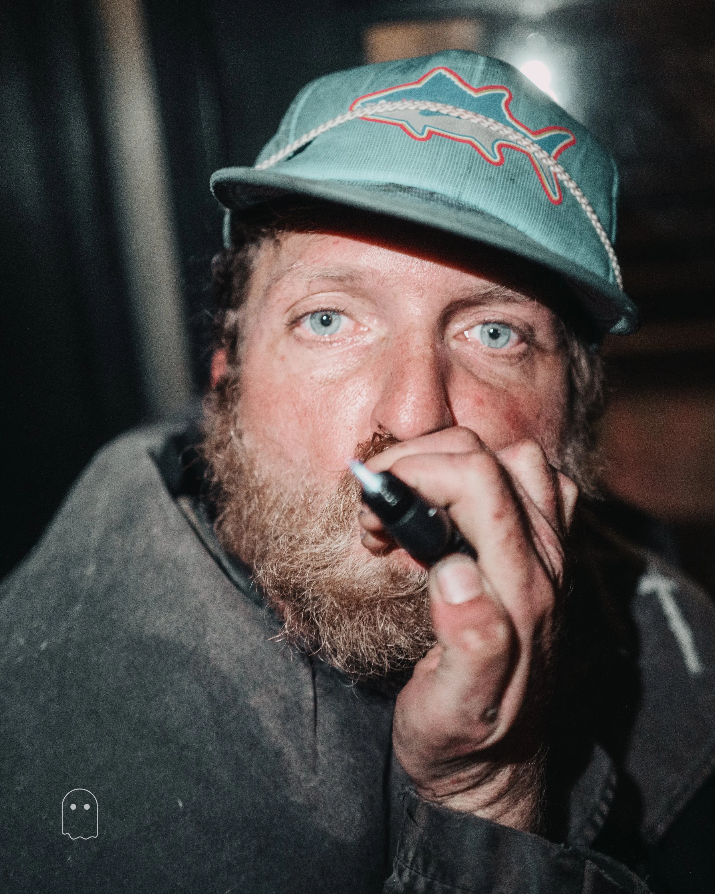 A man with a beard and blue eyes wearing a teal baseball cap with a Chevrolet logo and stitching, holding a black marker near his nose/pencil to his face, with dark background.