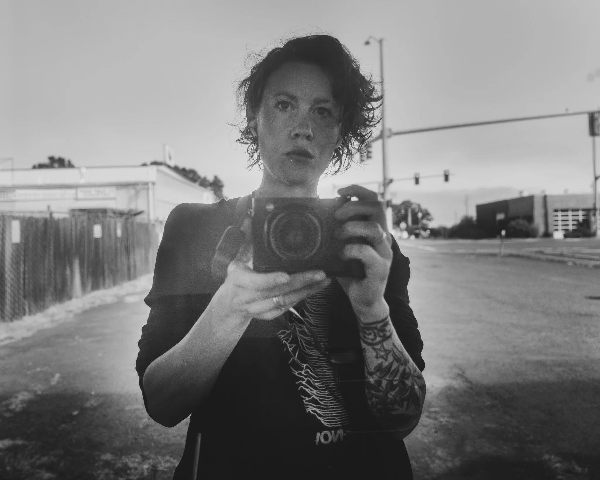 A woman taking a selfie in black and white, standing outdoors with traffic lights in the background. She has curly hair, tattoos on her arm, and is holding a camera or phone with both hands.