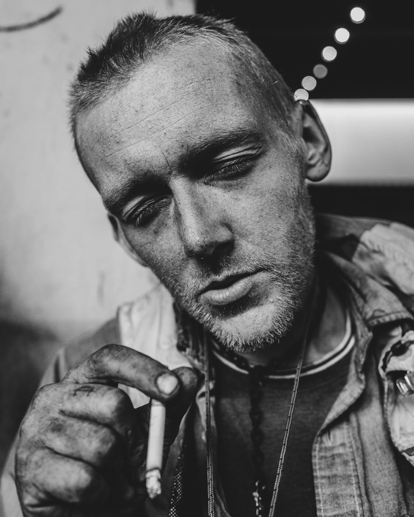 Close-up of a man with closed eyes, a serious expression, and short hair, holding a cigarette in his hand, wearing a jacket, in a black-and-white photograph.