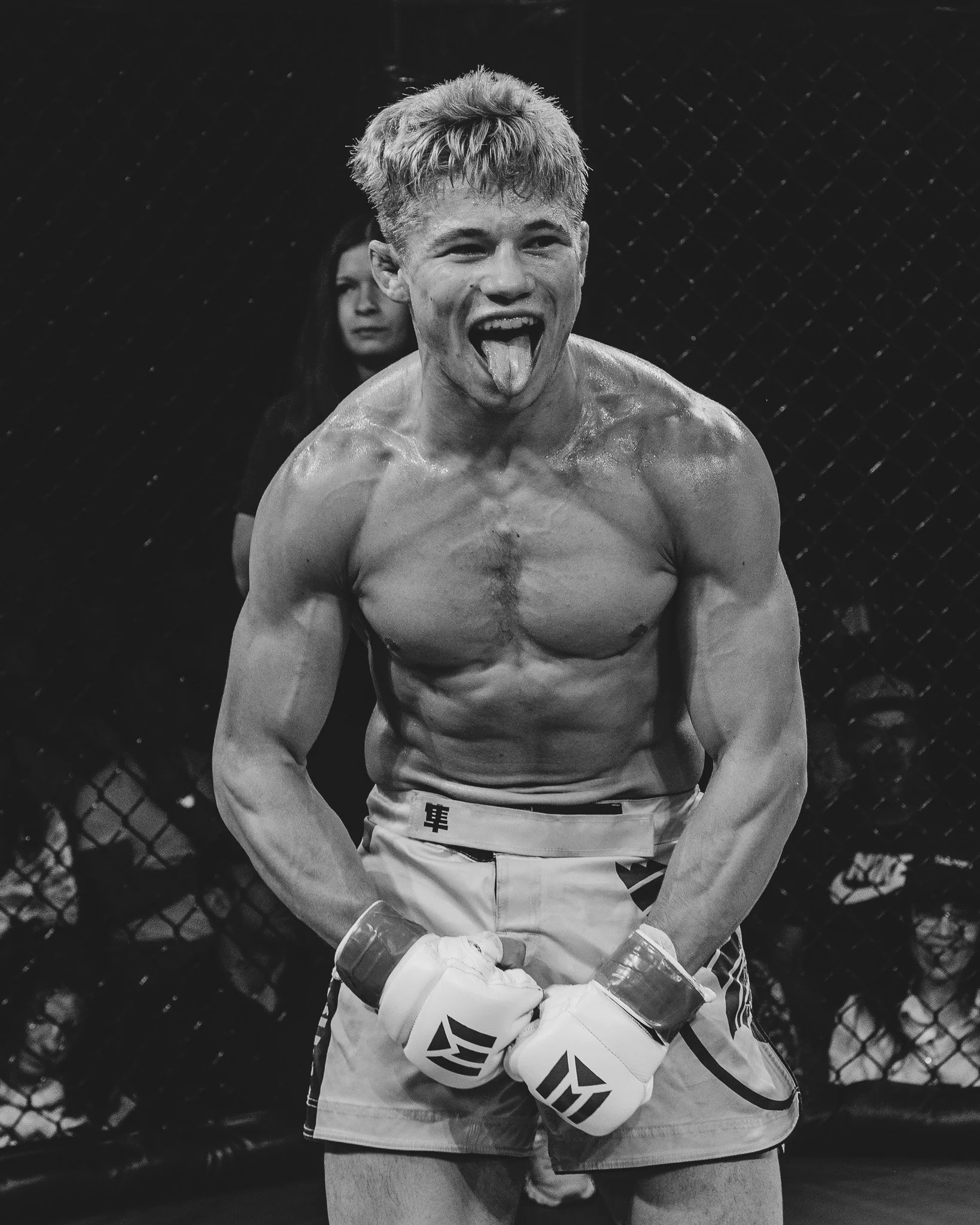 A shirtless male mixed martial arts fighter with blonde hair sticking out his tongue in a cage, with a serious female interviewer or trainer behind him, and a chain-link fence background.